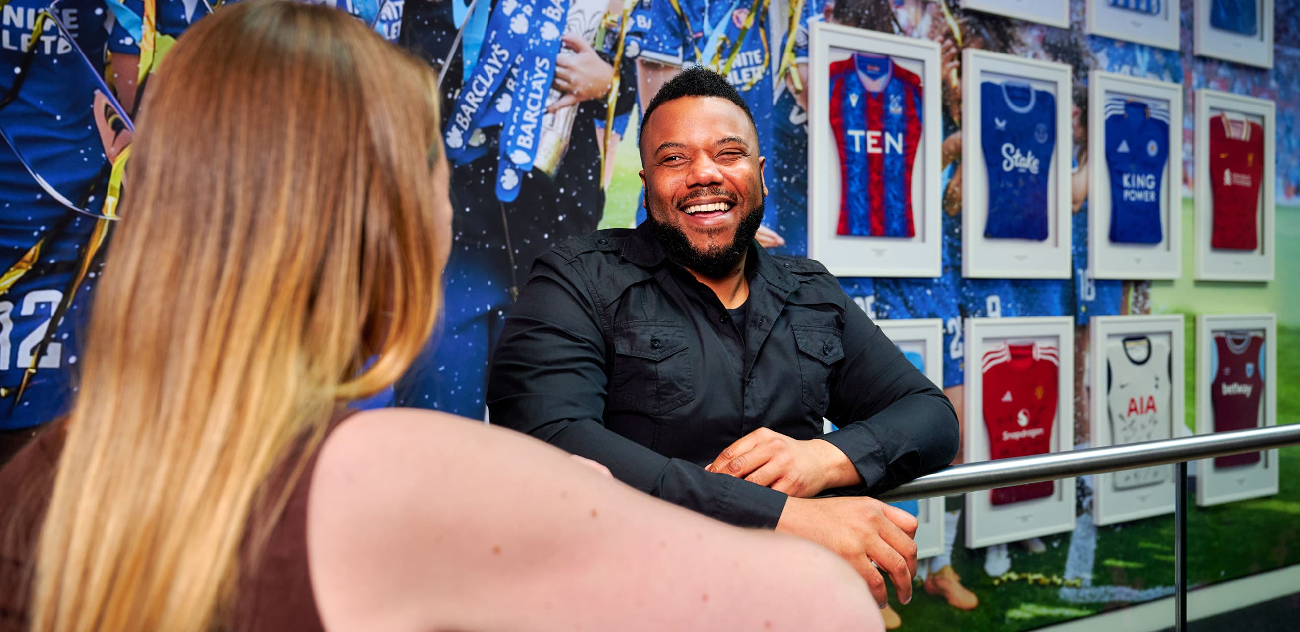 Man smiles while chatting with a colleague in front of framed football shirts on a wall.