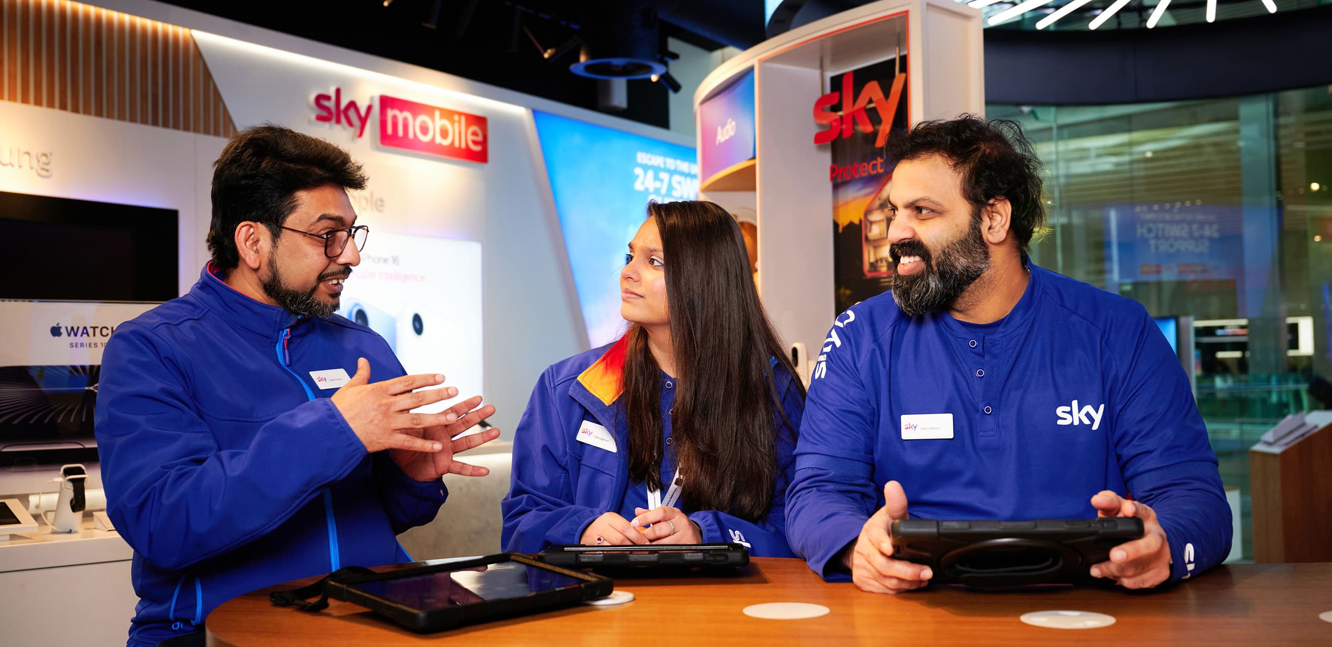 Three Sky employees in uniform discuss work at a table with tablets in a Sky retail store.