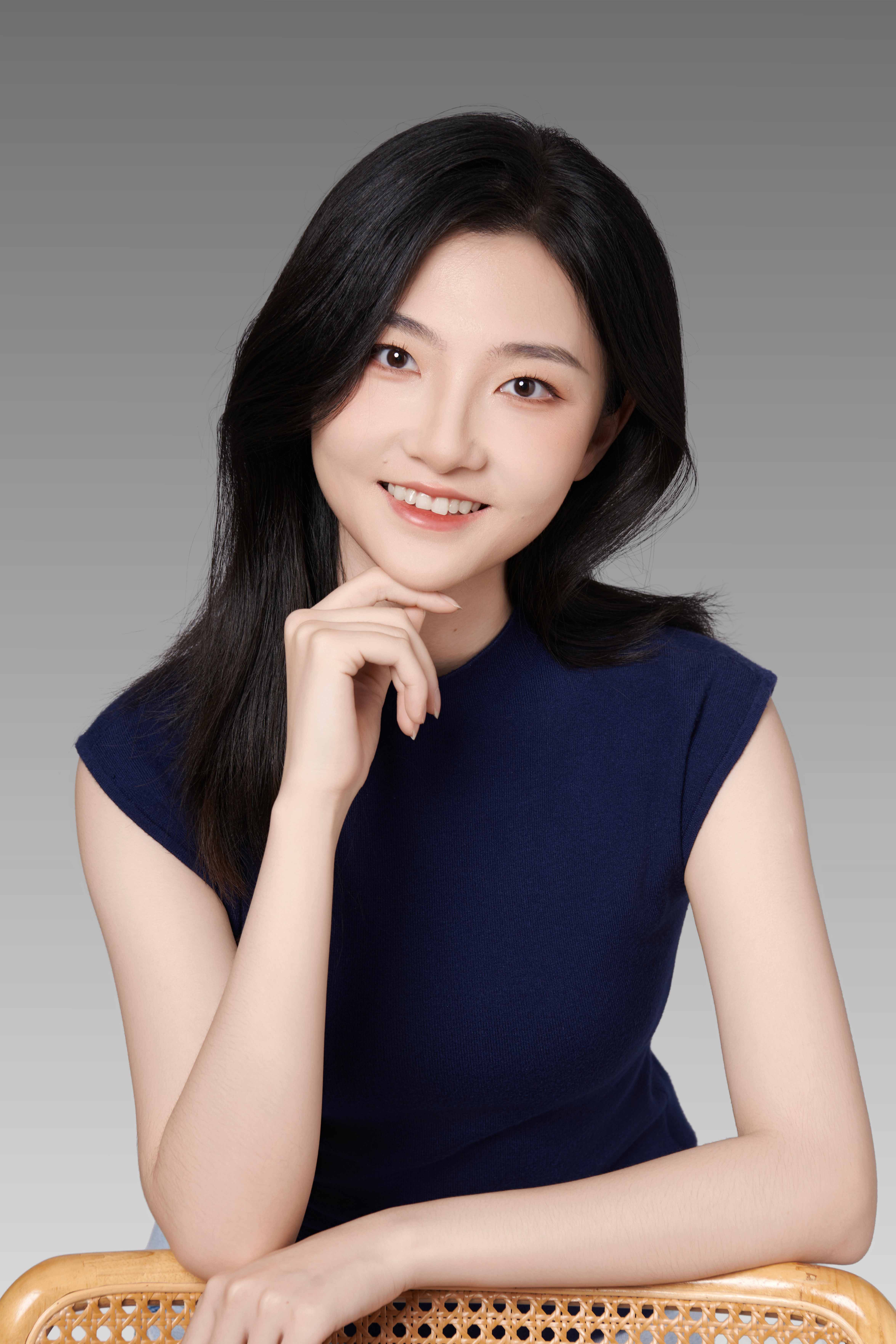 Wanxin is smiling posing leaning on a bamboo wooden chair infront of a white studio backdrop. She is wearing a nave blue sleavless dress with her right hand placed under her chin.