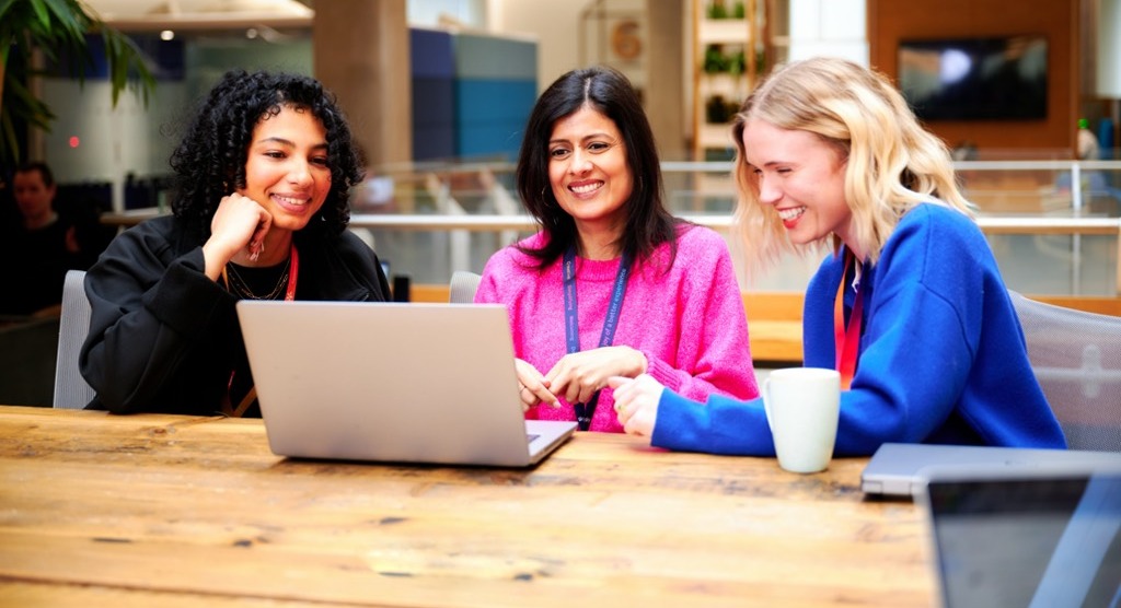 Three colleagues sit together at a large wooden table in an open‑plan Sky office, collaborating around a laptop. The workspace features exposed columns, modern lighting, indoor plants, and informal seating areas in the background, creating a bright and collaborative working environment.