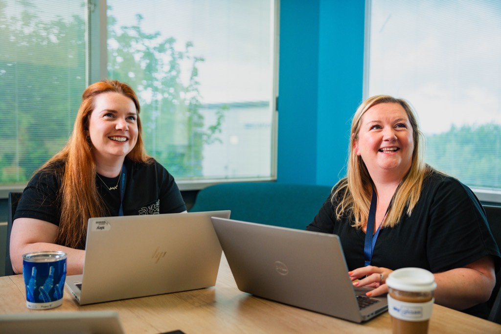 Two colleagues sit at a table in a bright meeting room with blue walls and large windows, each working on a laptop. A takeaway coffee cup and a reusable cup are placed on the table, and soft seating is visible in the background.