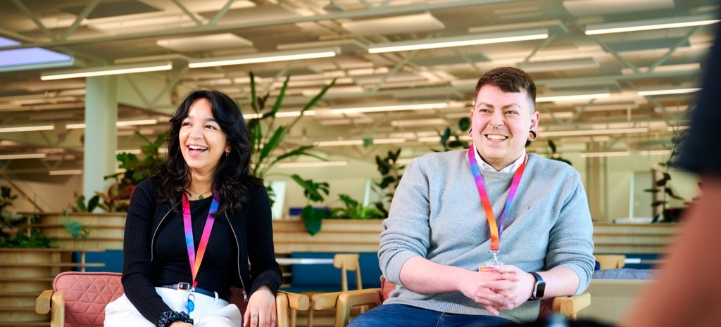 Two colleagues sit in lounge-style chairs in a bright, open office space with plants, wooden furniture and overhead lighting.