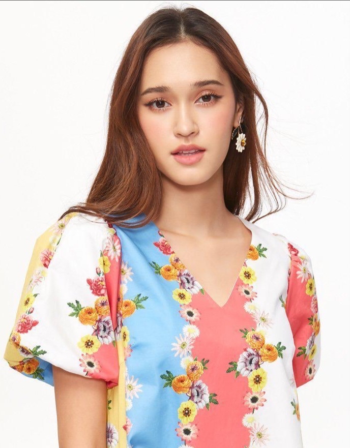 Emmy is wearing a floral short sleeve dress with her right shoulder forward posing intront of a white backdrop. She is also wearing a white daisy flower earring.