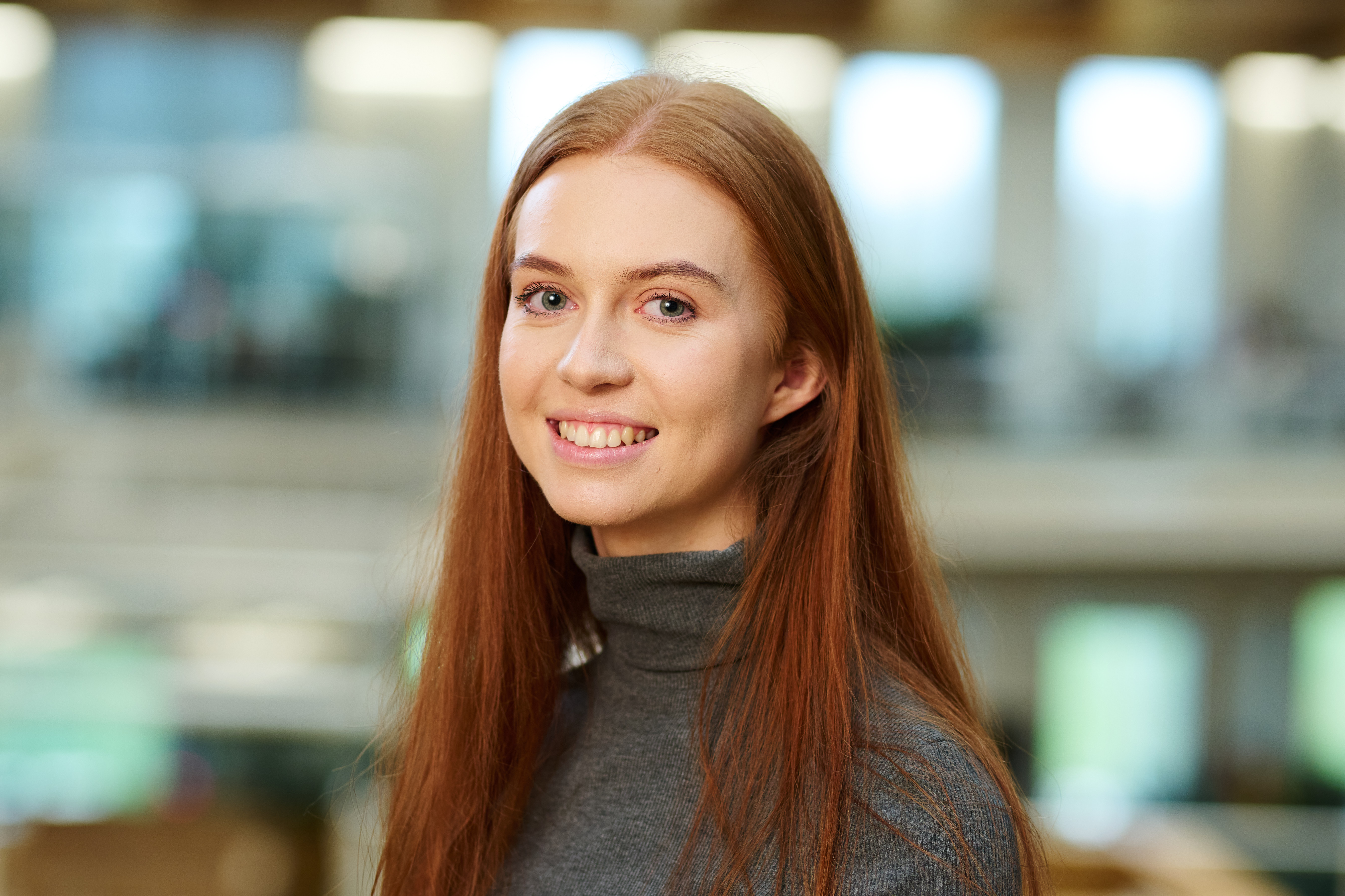 Rachel is wearing a grey turtle neck jumper with her hair down. smiling with the Sky office out of focus in the background
