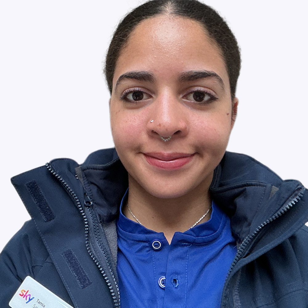 A close up portrait selfie of Tamia facing the camera against a plain white background. Tamia is wearing a dark padded Sky jacket over a blue collared Sky retail top with buttons. A Sky branded name badge is partially visible on the jacket, showing the Sky logo and the name “Tamia.” Tamia’s hair is tied back, is wearing small facial piercings. The image is tightly cropped around the head and shoulders, with even lighting and no background details.