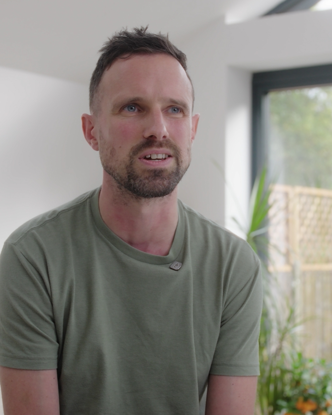 James is shown from the chest up in a softly lit indoor setting. James is wearing a plain, short sleeved green T shirt with a small clip on microphone attached near the neckline. They are positioned slightly angled to the left of the frame. The background includes a white wall, a large window with natural light coming through, and several green houseplants placed near the window. The depth of field is shallow, leaving the background slightly out of focus.