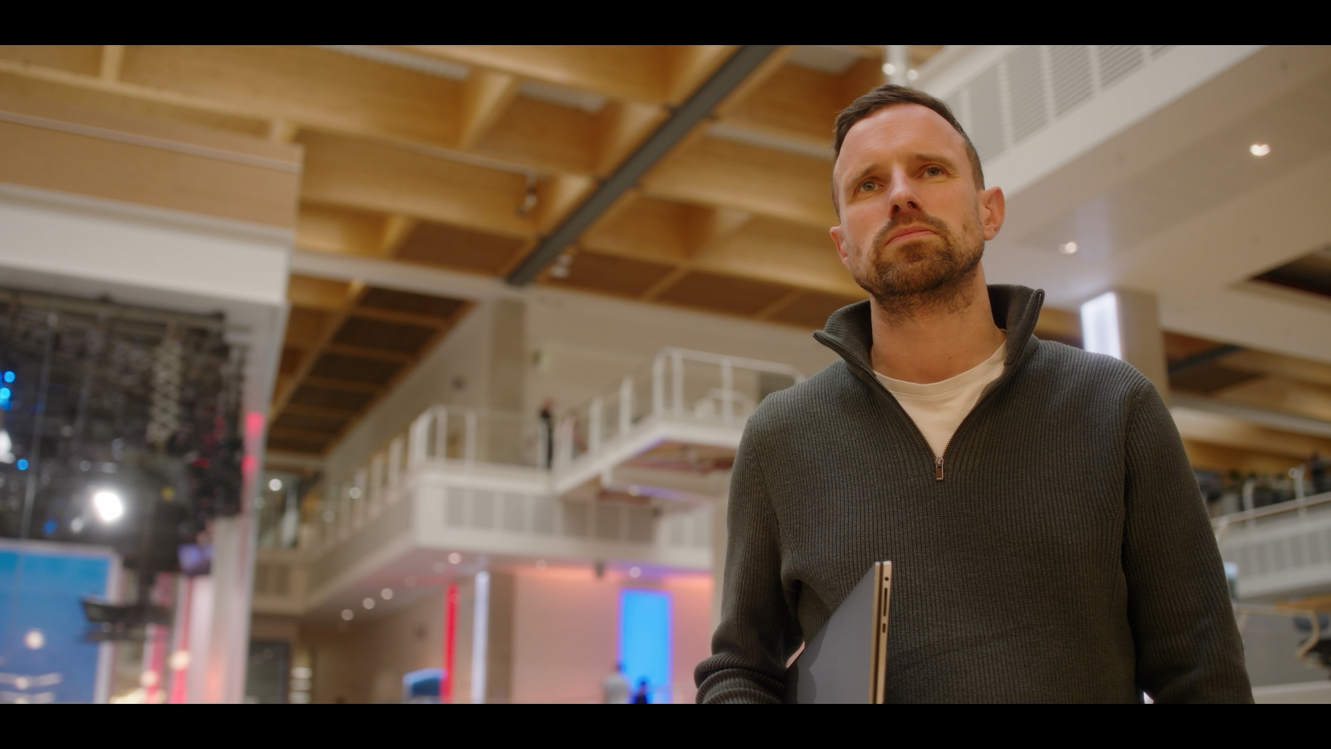 James is standing indoors in Sky Central, Osterley, with high ceilings made of light coloured wood panels and exposed beams. James is shown from the waist up, wearing a dark grey quarter zip sweater over a light coloured T shirt and holding a thin laptop or folder against their side. The background features the Sky News box, multiple open walkways with white railings, soft overhead lighting, and blurred coloured light panels in blue and pink tones.