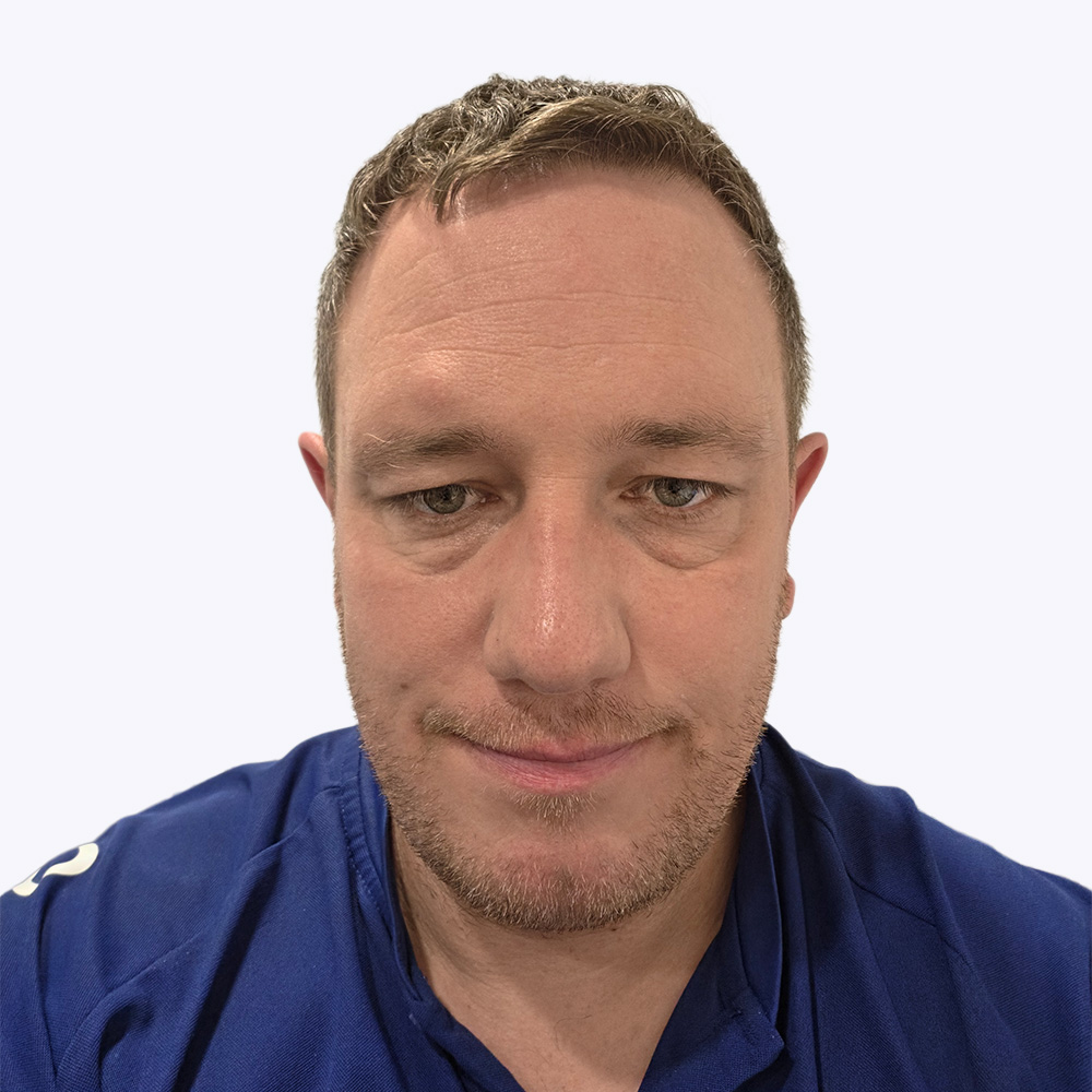 A close‑up head‑and‑shoulders photograph of a person facing the camera against a plain white background. The person has short hair and light facial stubble around the jaw and upper lip. They are wearing a blue collared top with visible stitching near the neckline and shoulders. The image is tightly cropped, with even lighting and no visible background objects or text.