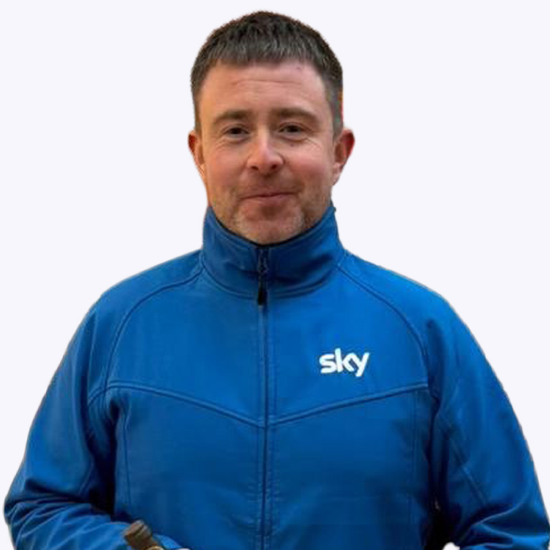 A head‑and‑torso photograph of a Oliver standing against a plain white background. Oliver is wearing a Sky-branded blue zip‑up jacket with a high collar zipped to the neck. The Sky logo is printed in white on the left side of the chest. The jacket has visible seam detailing across the front. The image is evenly lit, with no background objects or additional text visible.