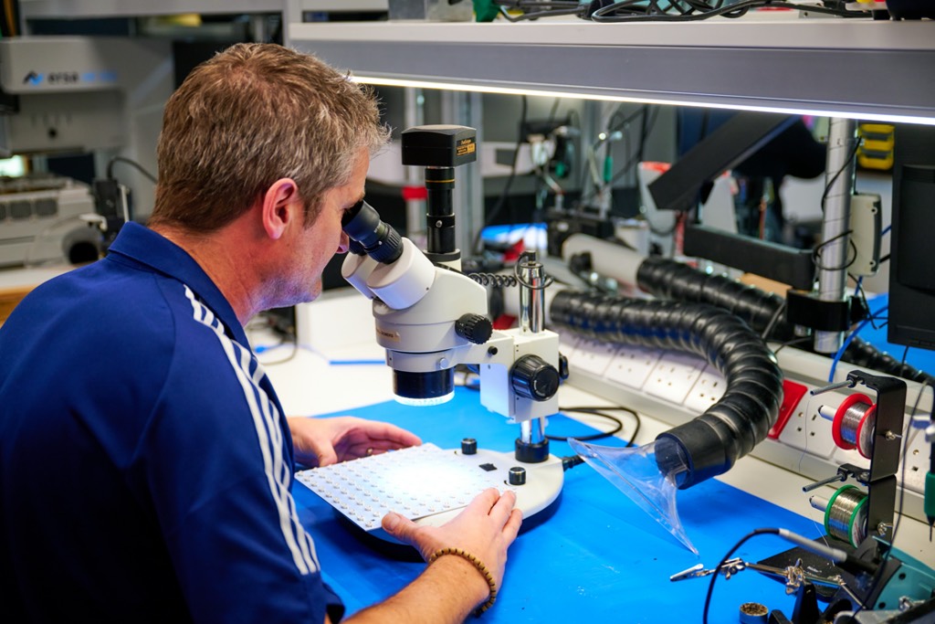 A technician works at an electronics workstation, using a microscope to examine a small component on a circuit board. The desk is equipped with tools, wires, and ventilation tubing in a well-lit, modern lab environment.