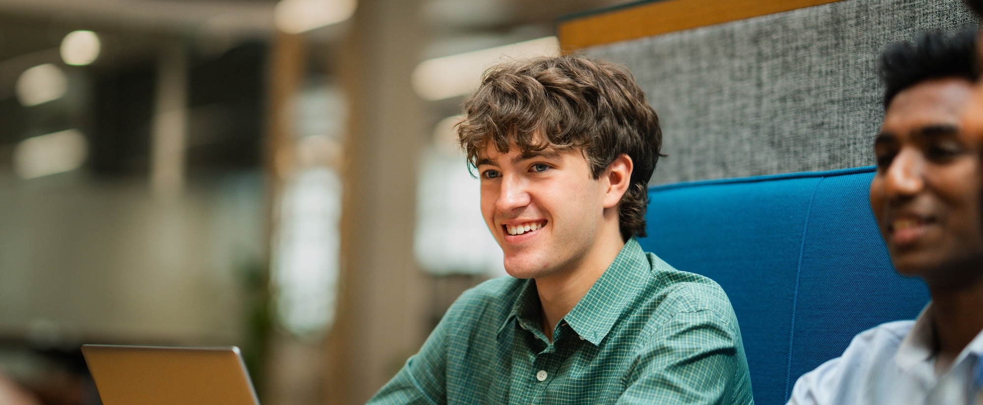 Smiling man in a green checked shirt using a laptop at a shared office table, with colleagues seated beside him.