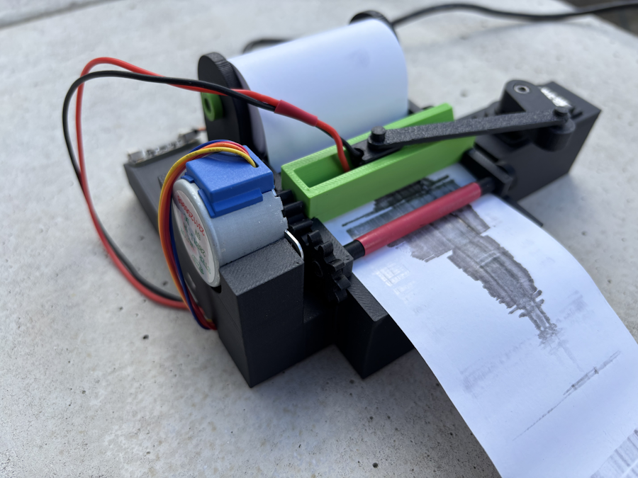 The device is 3d printed plastic in green and black. There is a roll of white paper that is marked with a black seismographic like image.