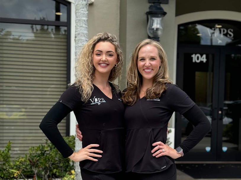 Two smiling staff members wearing black scrubs stand side by side with their arms around each other in front of VIPS.