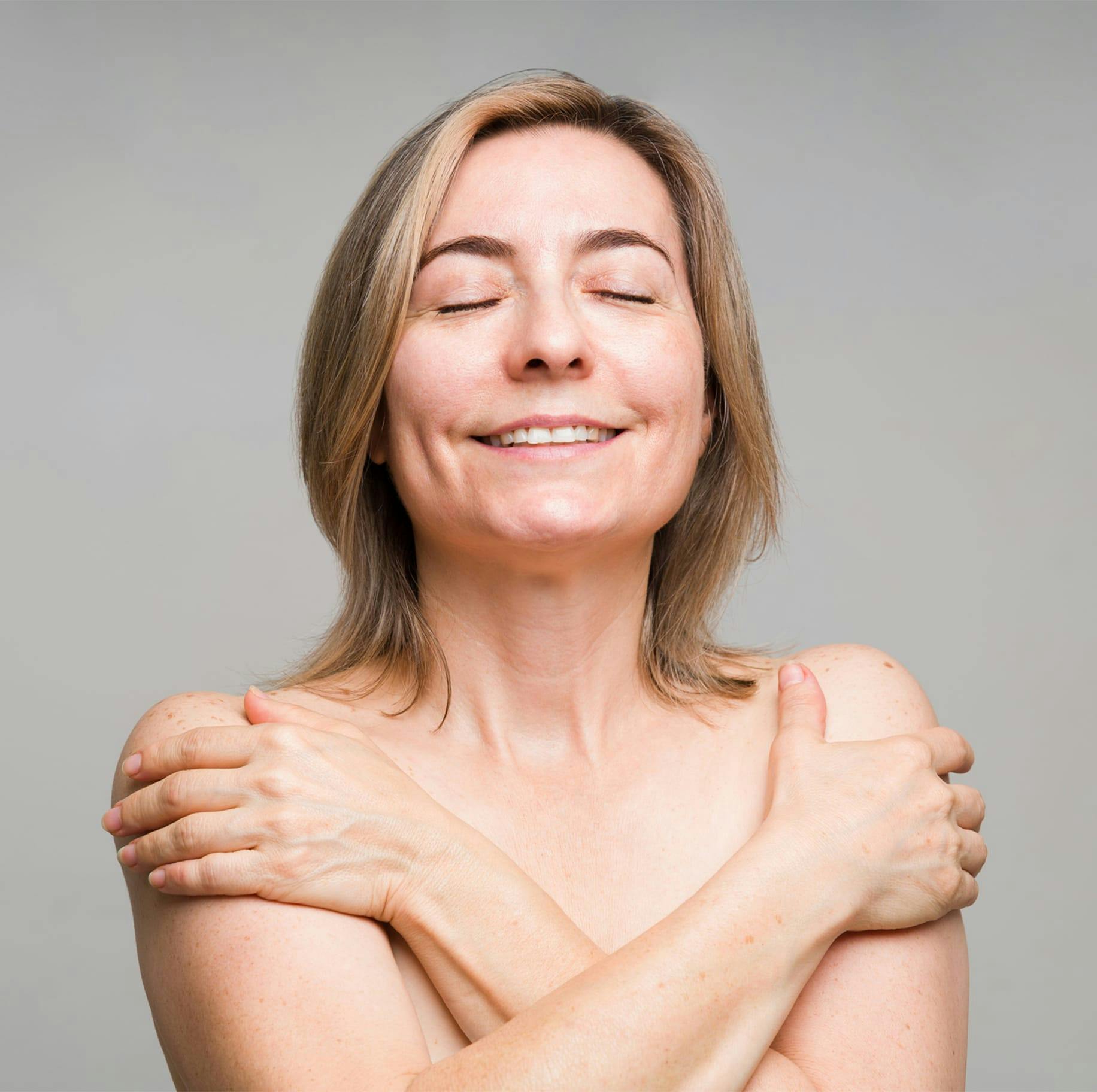 smiling woman with no shirt on crossing her arms to cover her breasts