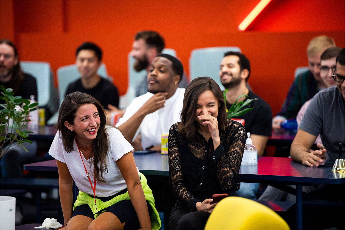 Two female colleagues in focus in the front frame laughing during a large team meeting