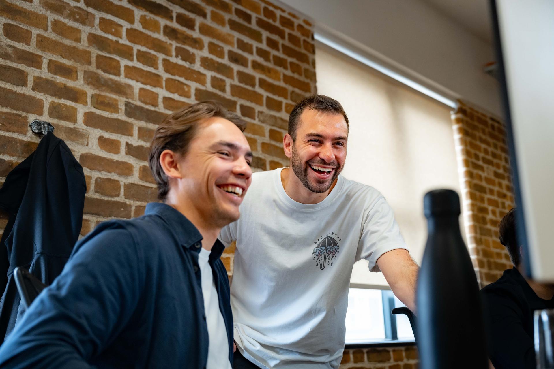 Male colleagues laughing at something on a computer screen in a bright modern office with brick walls