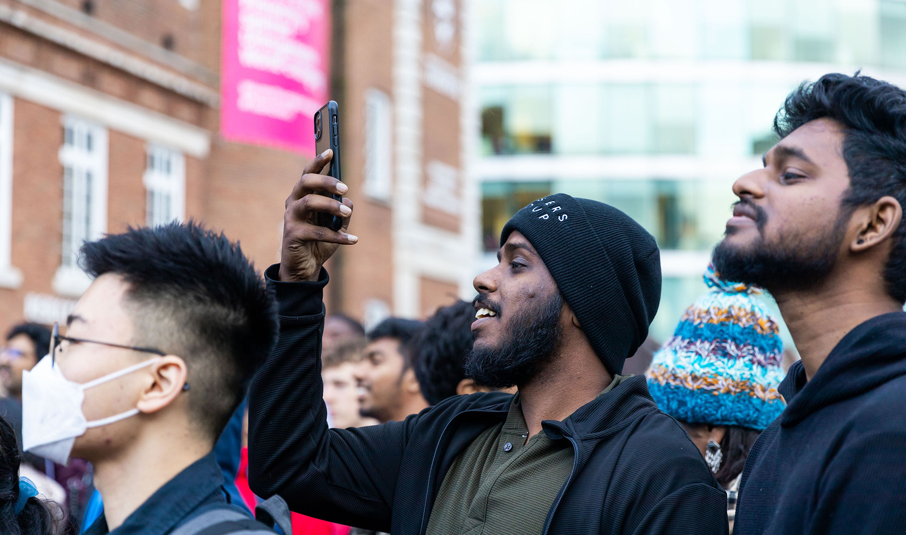 Crowd of male students craning their necks to looks for a winner. One student is filming on his iPhone.