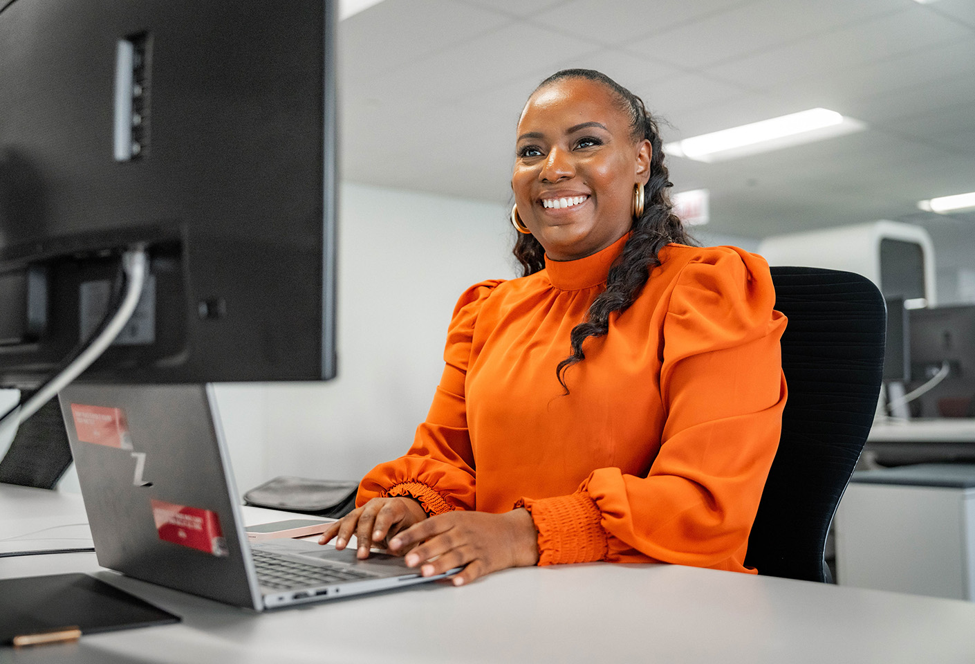 Image of woman wearing a vibrant orange blouse in an office, working at a desk with a laptop and a large screen