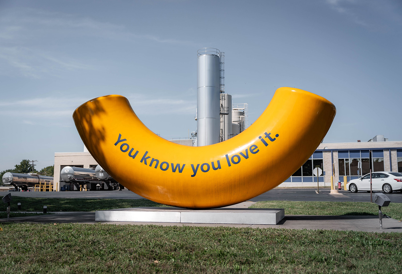 Image of huge macaroni sculpture outside of a factory, the macaroni has "you know you love it" printed on it