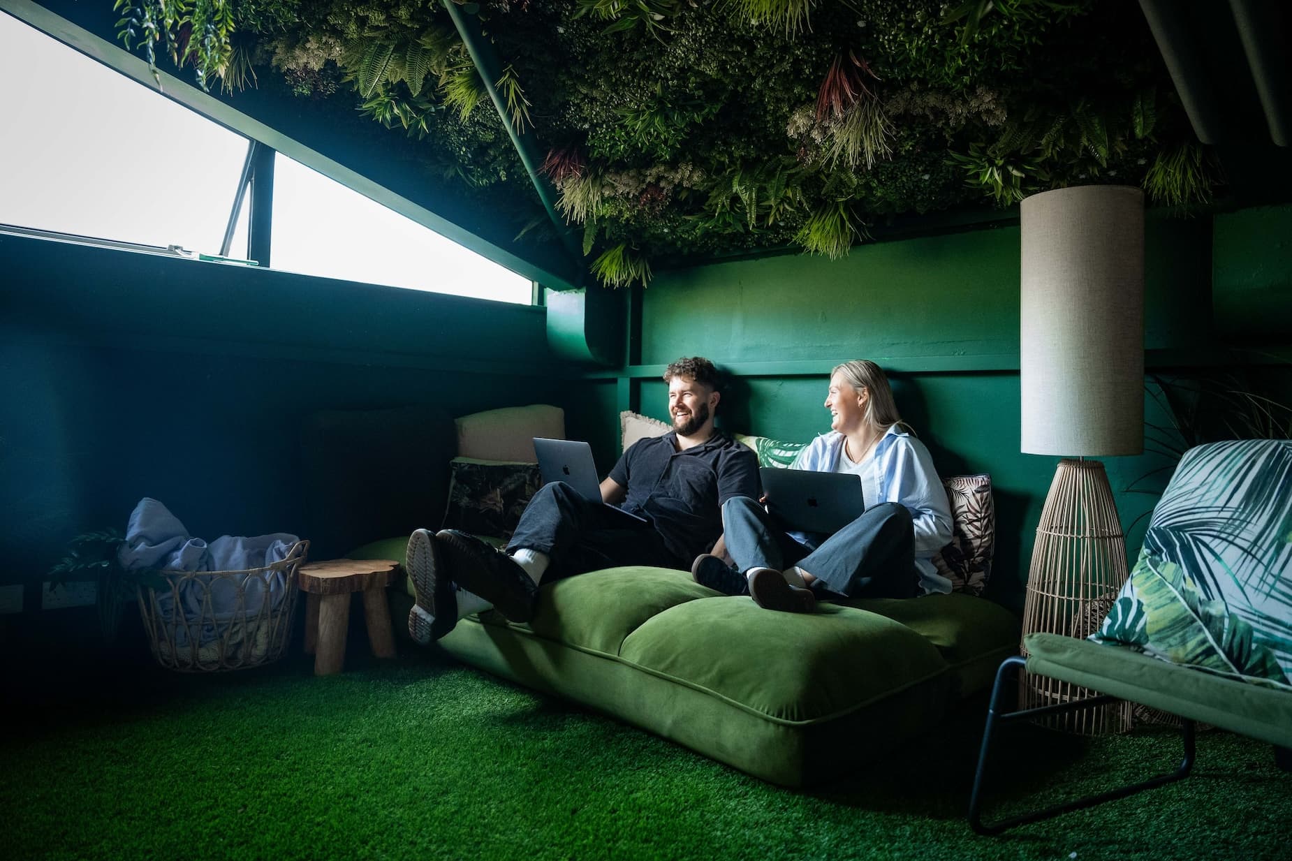Two wiser employees sit in a rainforest themed space on a green couch.