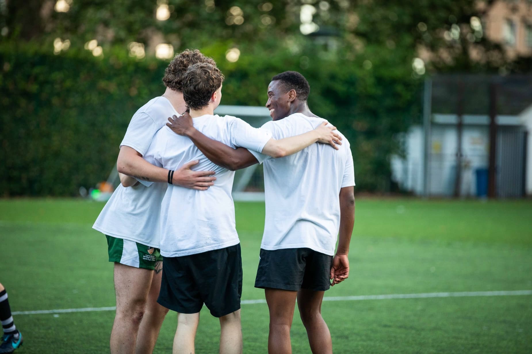 Three males stand on a football pitch with their arms round each other in gym gear.