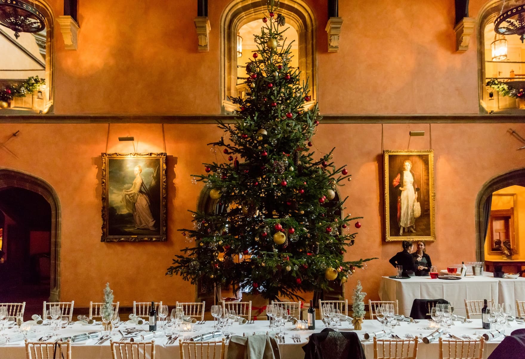 In a large dinner room, a Christmas tree stands in between paintings on the wall.
