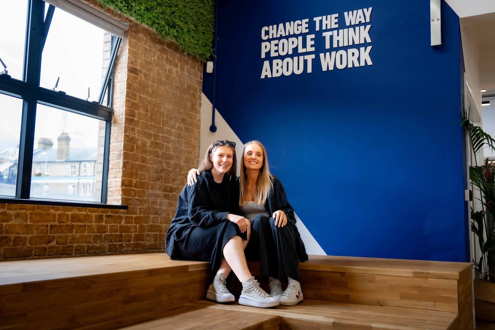 Two females with their arm round each other sit in the Wiser office smiling.
