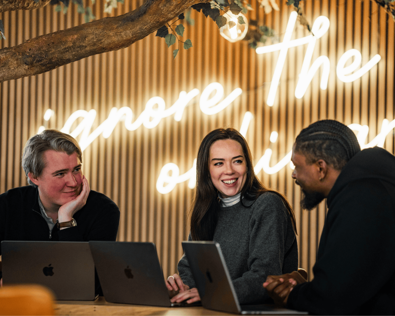 Two males and one female smiling in a meeting at a table with a neon sign saying ignore the ordinary behind them