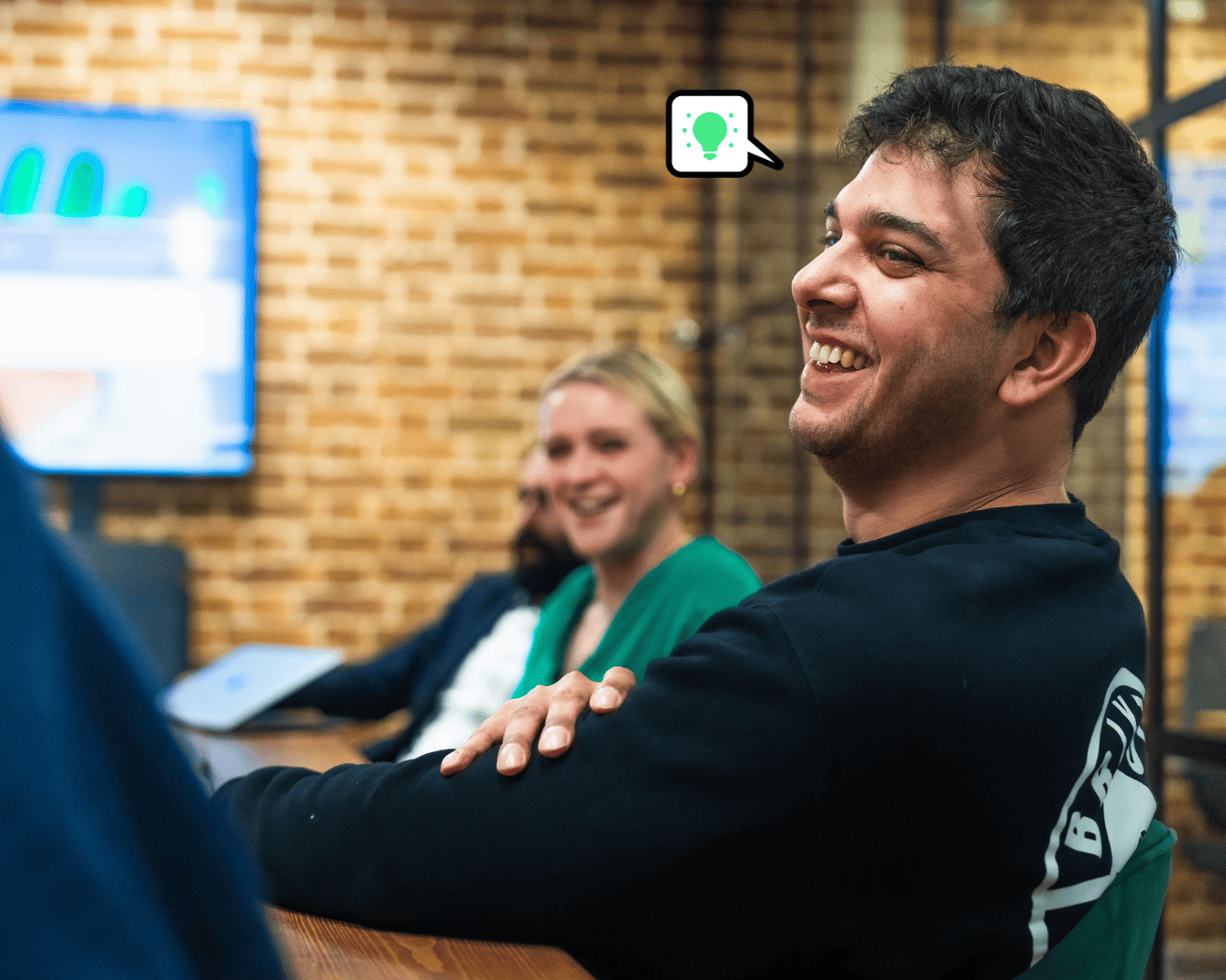 Photo of brown haired male in a black long sleeve top in a meeting with a female in a green top blurry at next to him