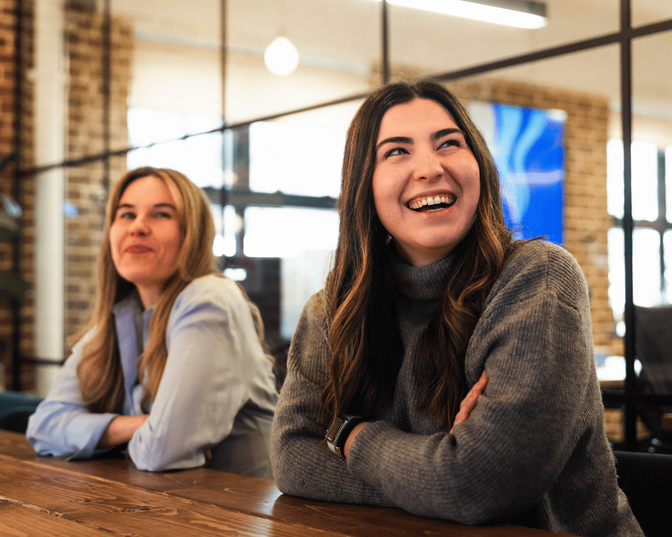 blonde female in a blue shirt and brunette female in a grey jumper in a meeting laughing looking at a screen