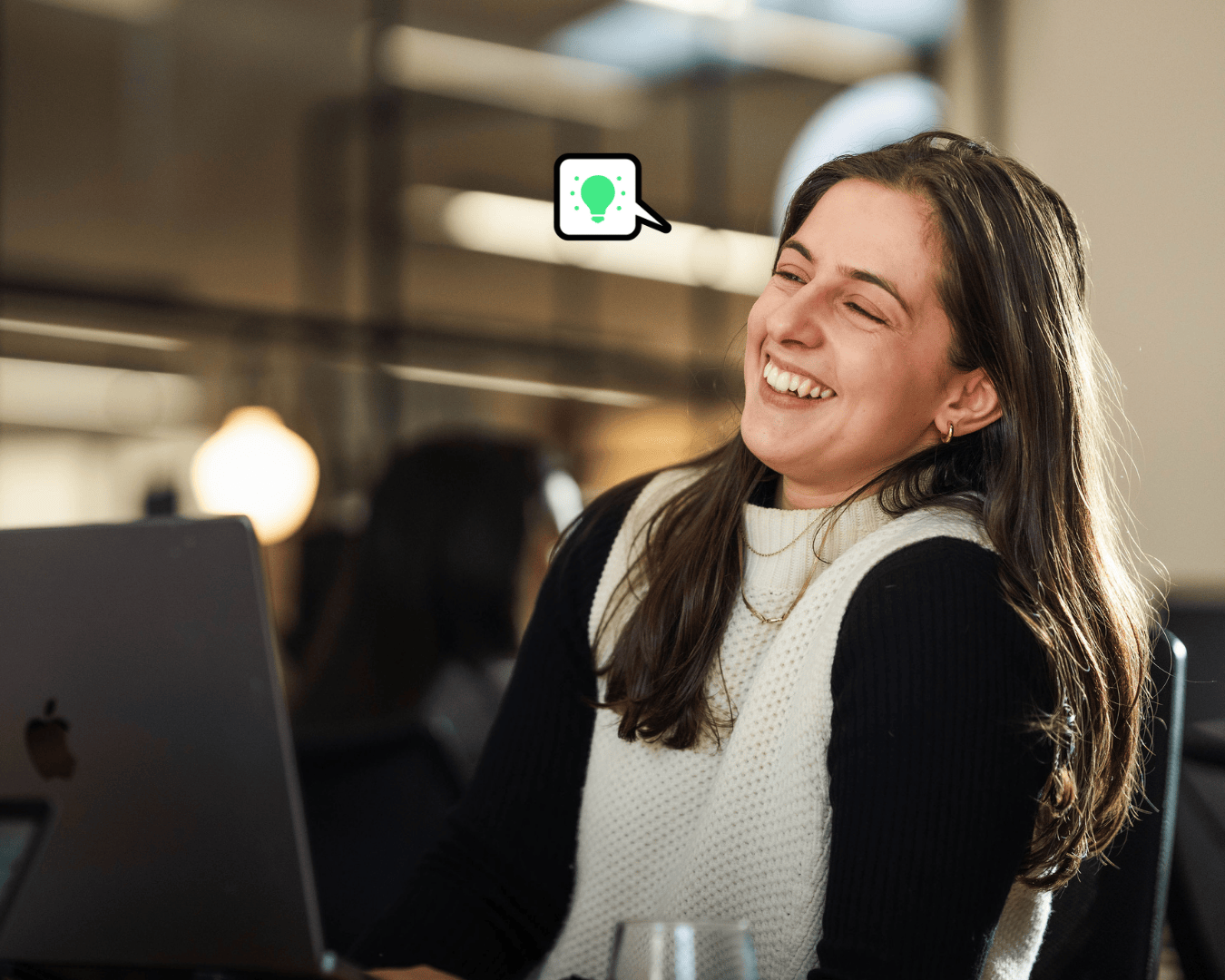 Photo of brown haired woman laughing at her desk with a laptop in front of her. In a long sleeve black top and white knitted vest
