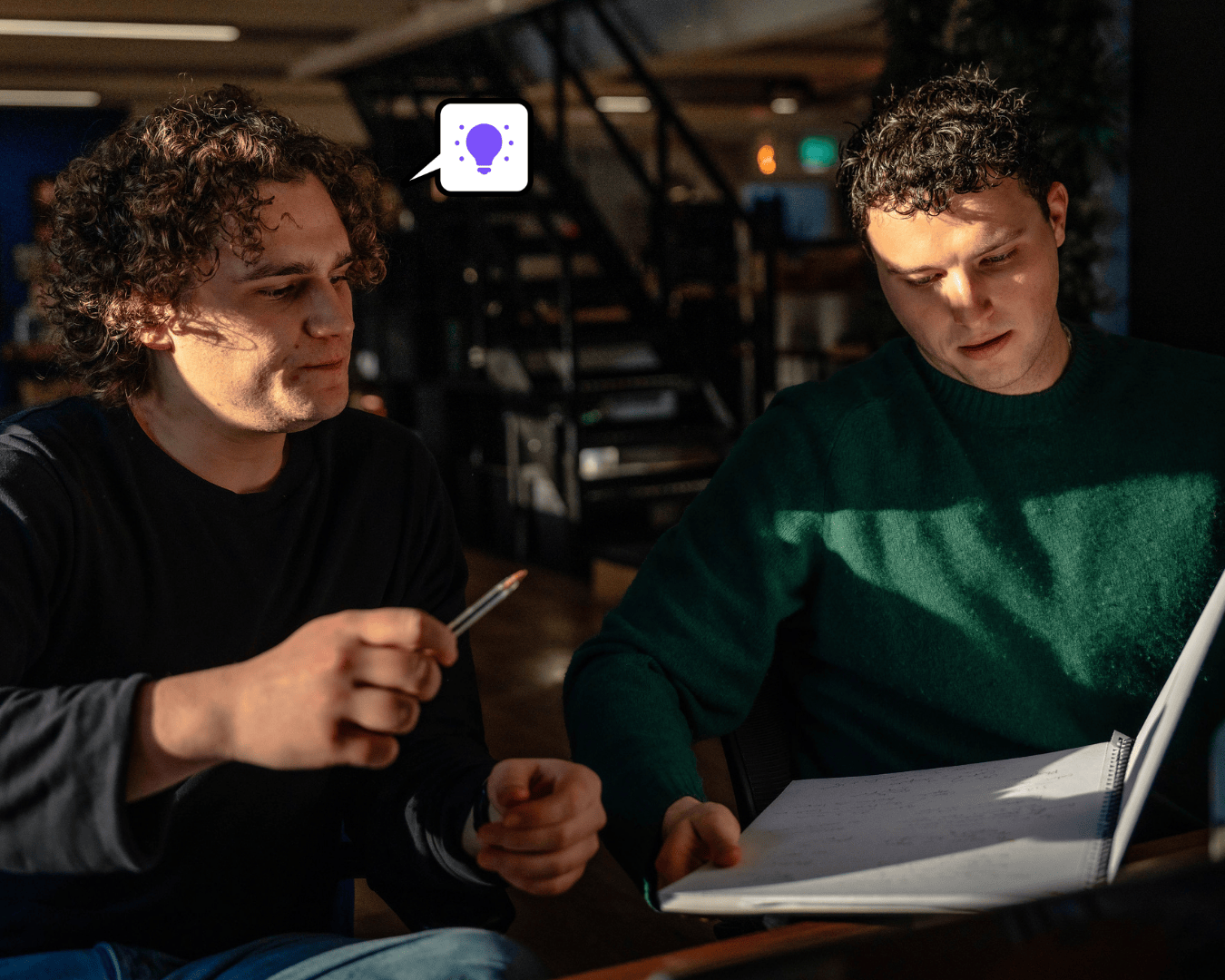 Photo of two males with brown hair looking at a large notebook one is in a green jumper the other in a black jumper