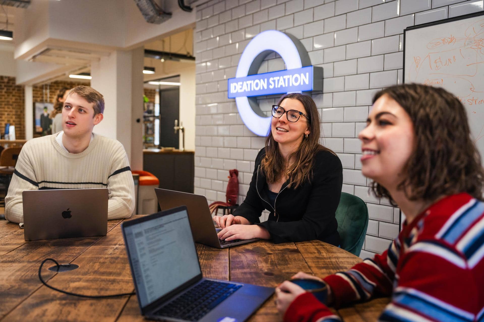 Image of three colleagues looking up at a screen, they have their laptops open and are smiling at what they are seeing