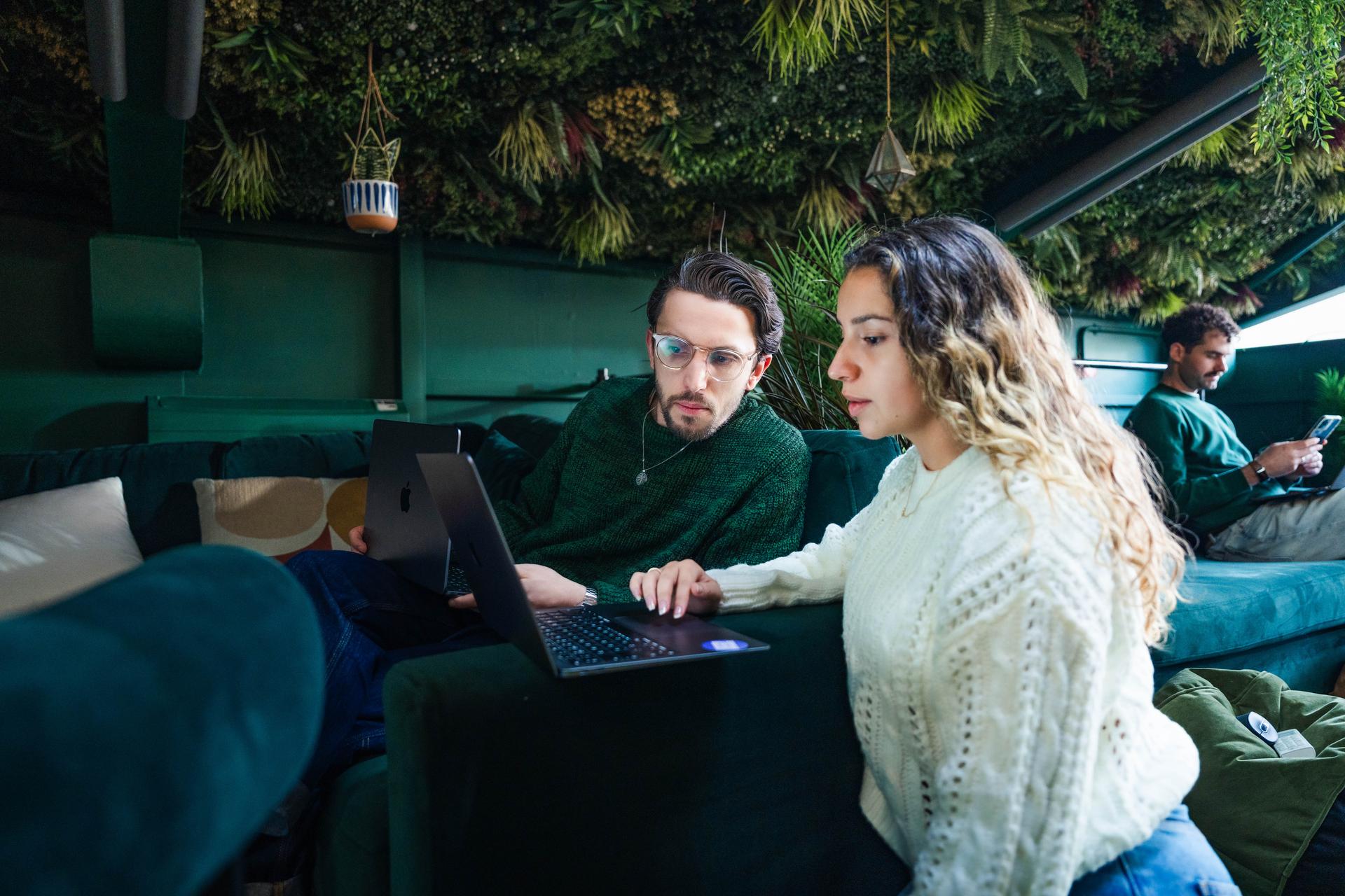 Two colleagues work in a green room with lots of faux foliage. The man is in his thirties, is sat on a green sofa, the woman is in her twenties, and is crouched near him, showing him something on her laptop