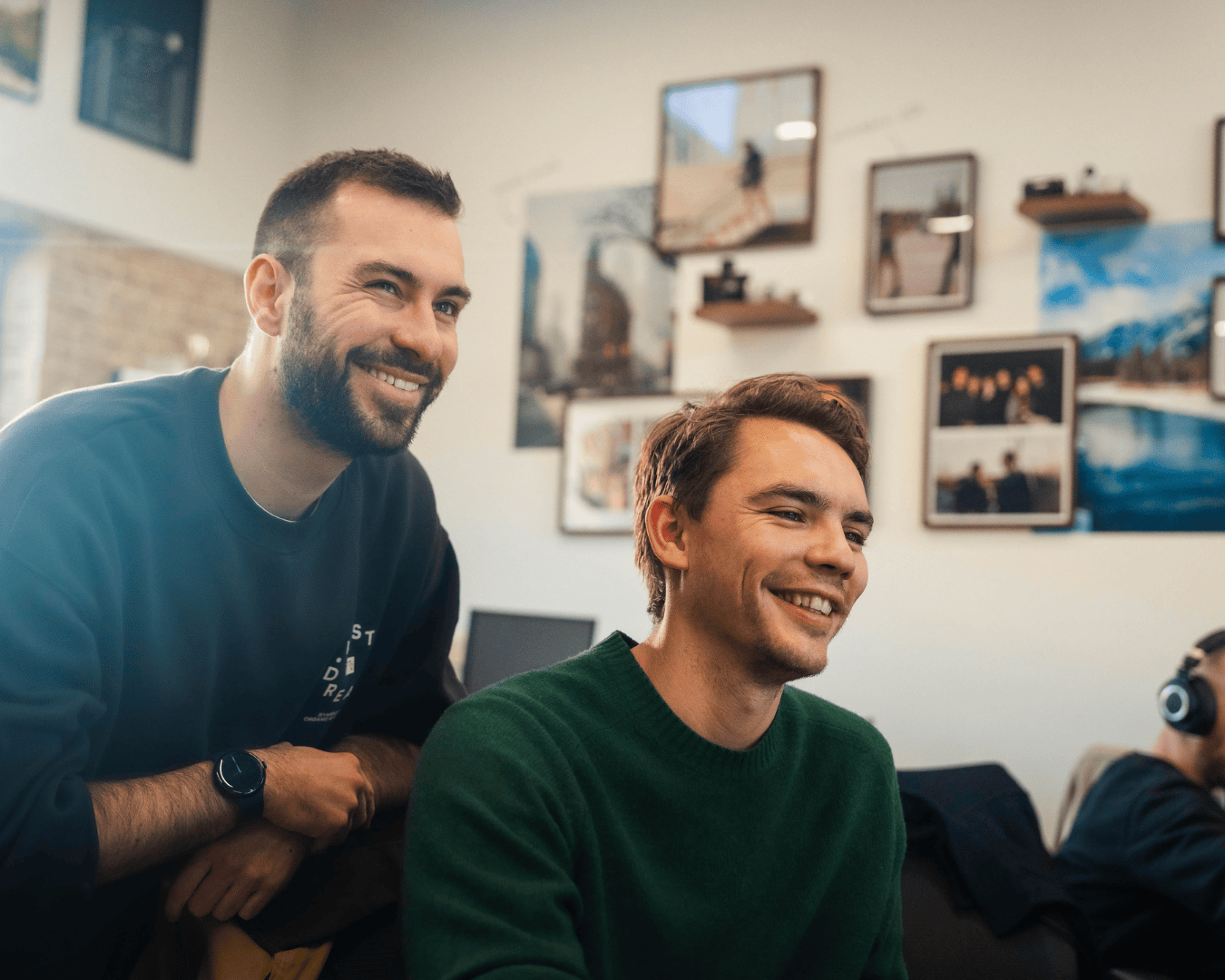 Photo of two males smiling at a screen with a photo frame wall behind them