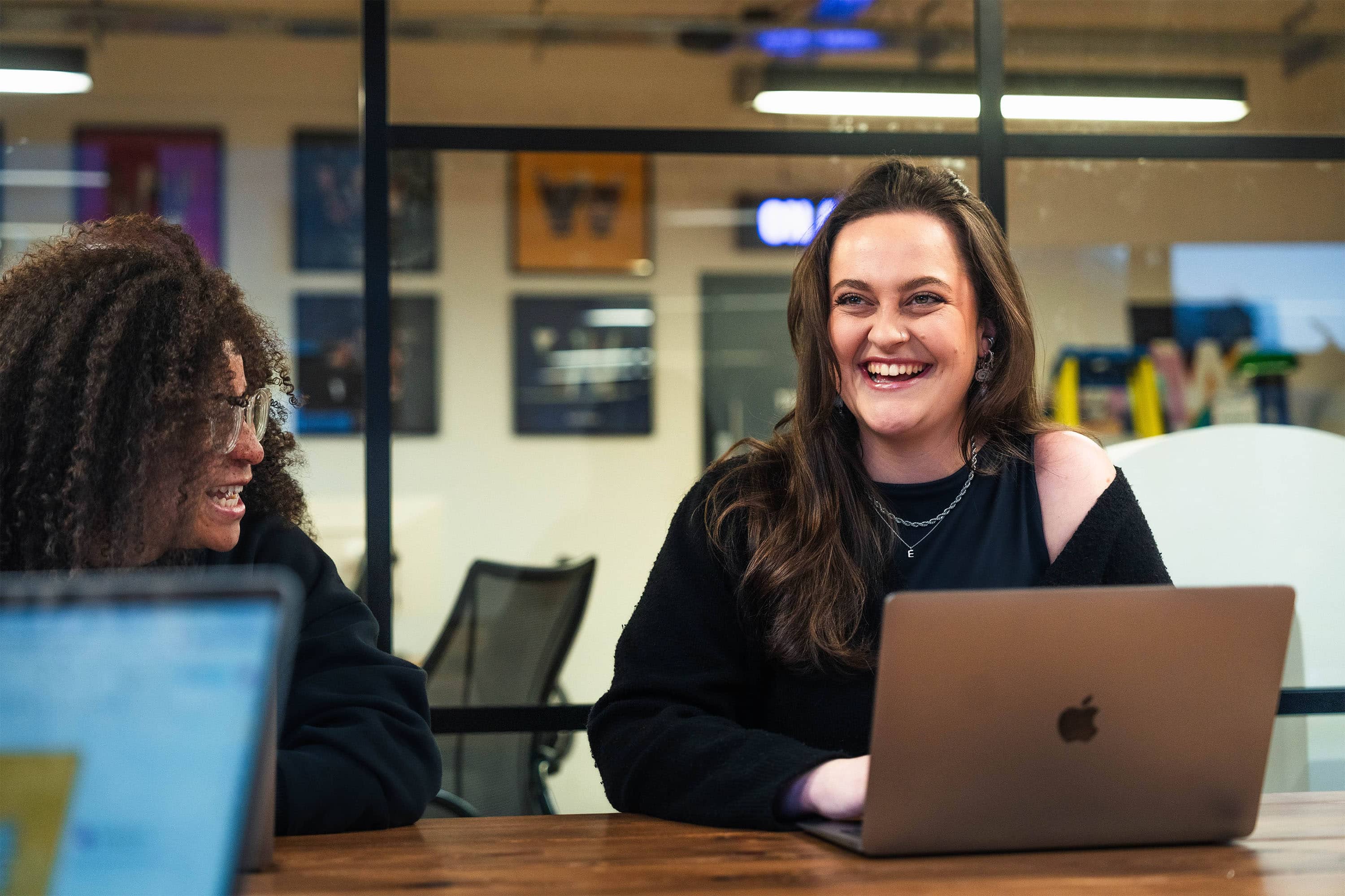 Two colleagues laugh during a meeting. They're both female, both young adults, one is white with long brown hair, one is black with long textured hair