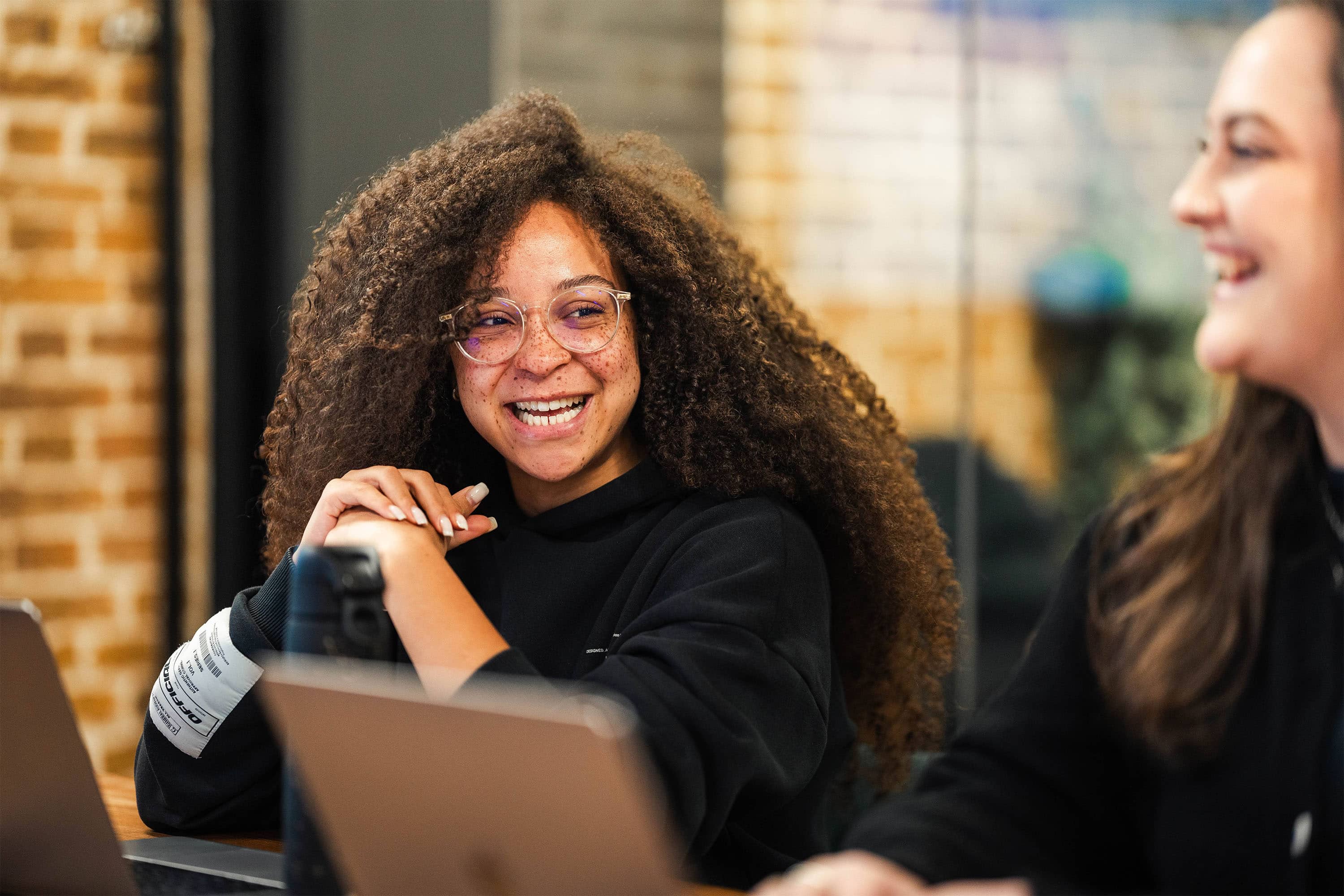 Young black adult woman smiles during a meeting, she has lots of freckles, is wearing glasses, and has long textured hair