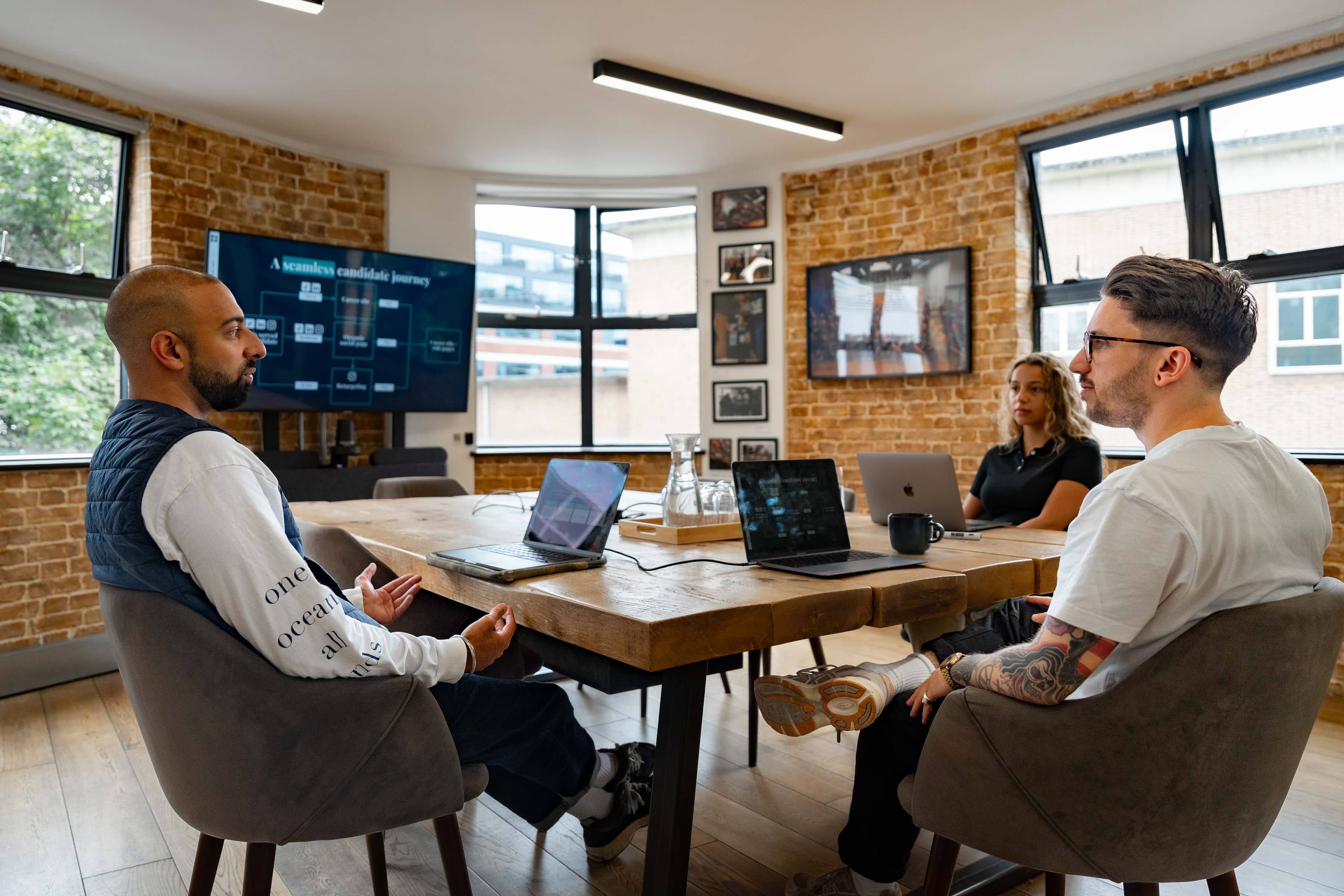 Three colleagues sat together in a meeting room discussing a presentation deck on their laptops. One man is indian, wearing a navy quilted vest, another man is white, with tattoos and brown hair, wearing a white t-shirt, the third person is a mixed race woman wearing a black t-shirt