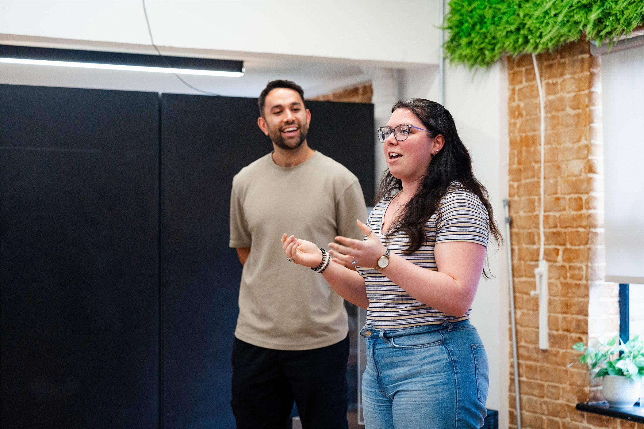 Two people are giving a presentation in the light bright Wiser office. One is a mixed race man in his thirties, wearing a beige brown t-shirt, the other is a white woman with long very dark hair, wearing glasses, a striped t-shirt and blue jeans
