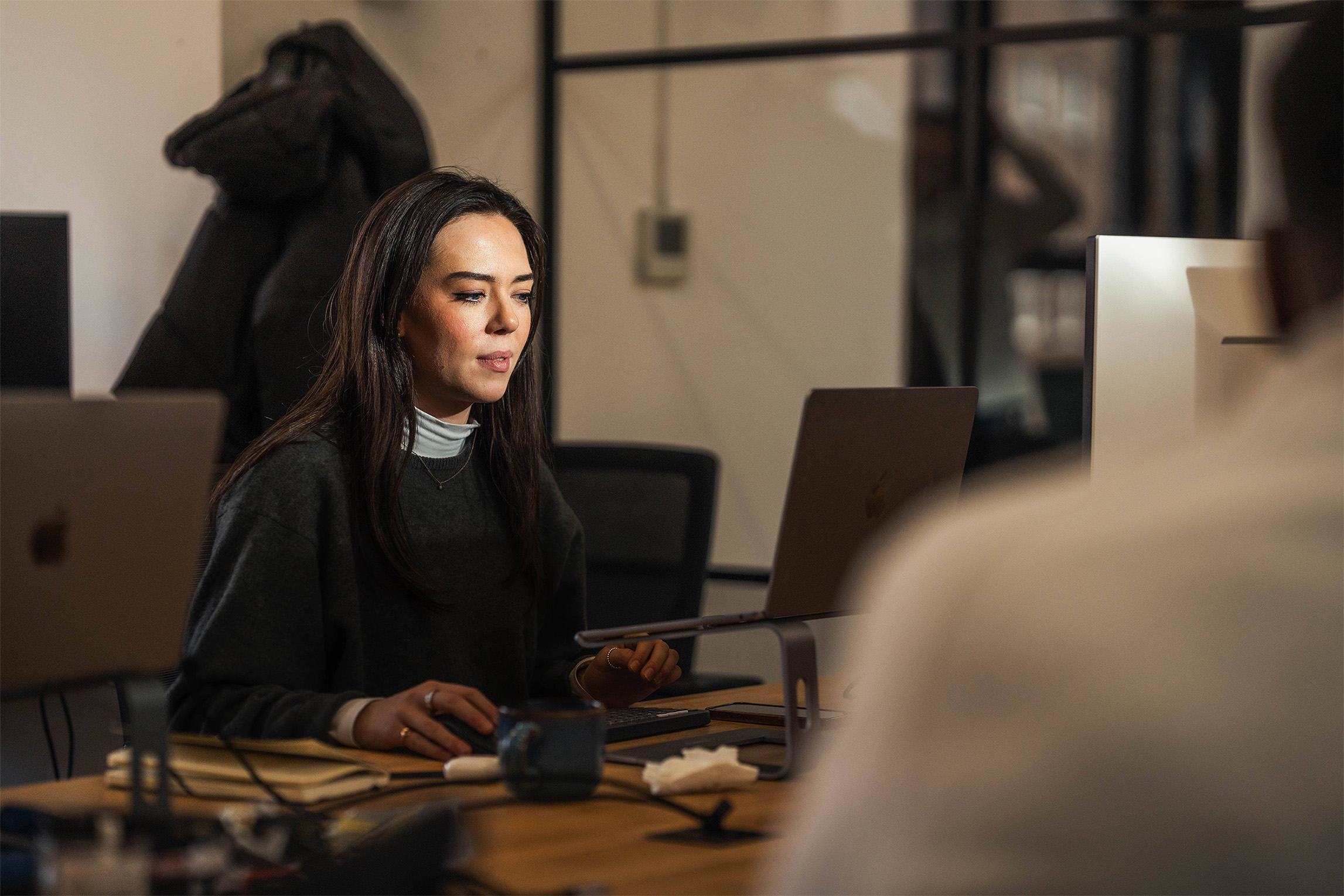 White woman in her twenties with long very dark hair works at her desk using a mouse and laptop stand for her laptop