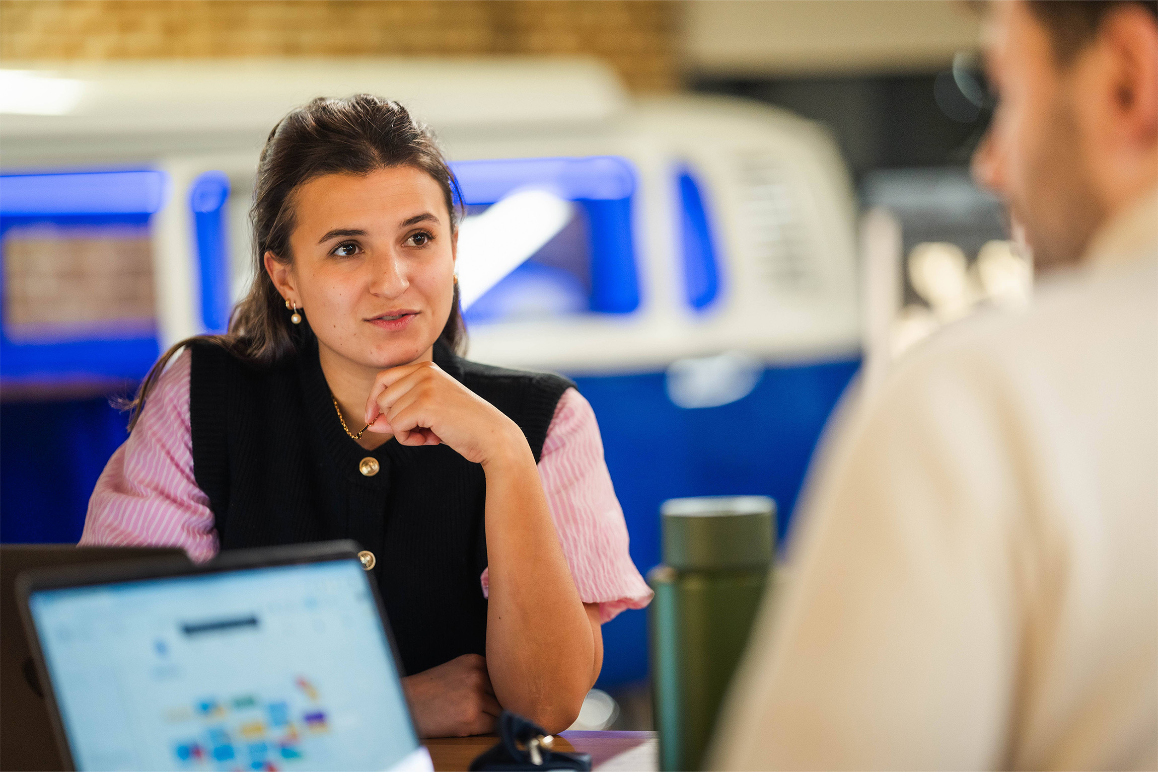 Young mixed race woman with dark hair, she's wearing a pink short sleeved shirt with a navy knitted cardigan over the top, she's having a conversation with another colleague, who is just out of frame