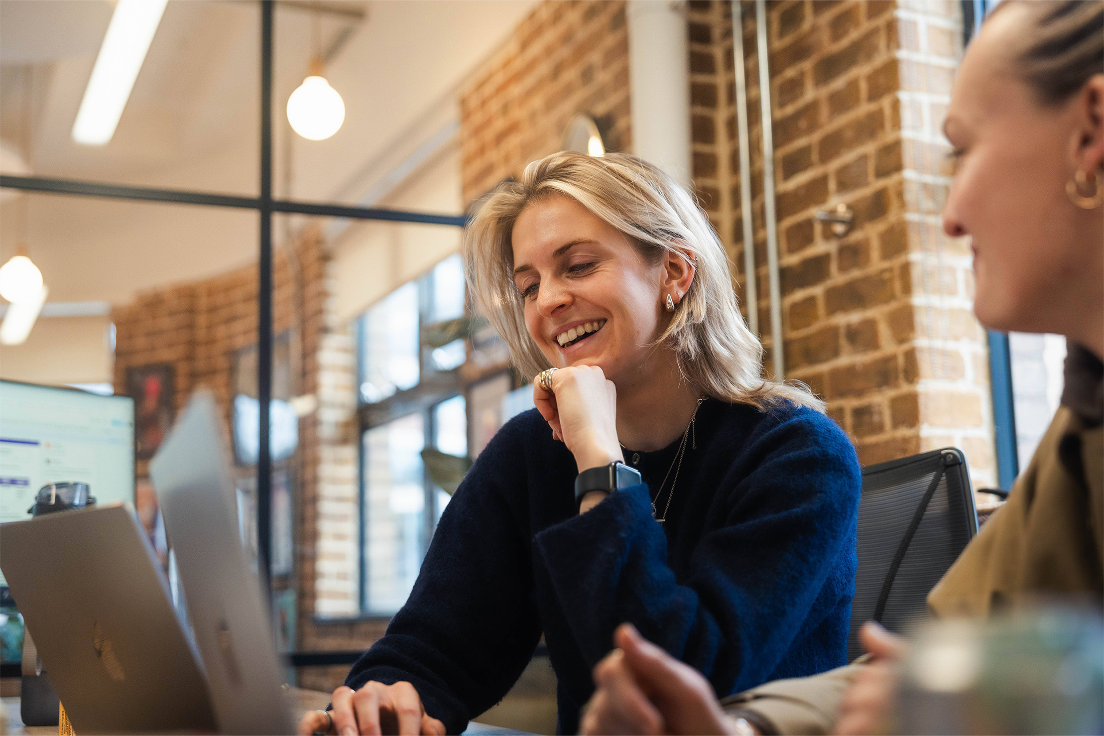Two women sat at a desk are looking at one laptop together. They're both blonde, and the women who's the centre of the shot is wearing a dark navy knitted jumper and an apple watch