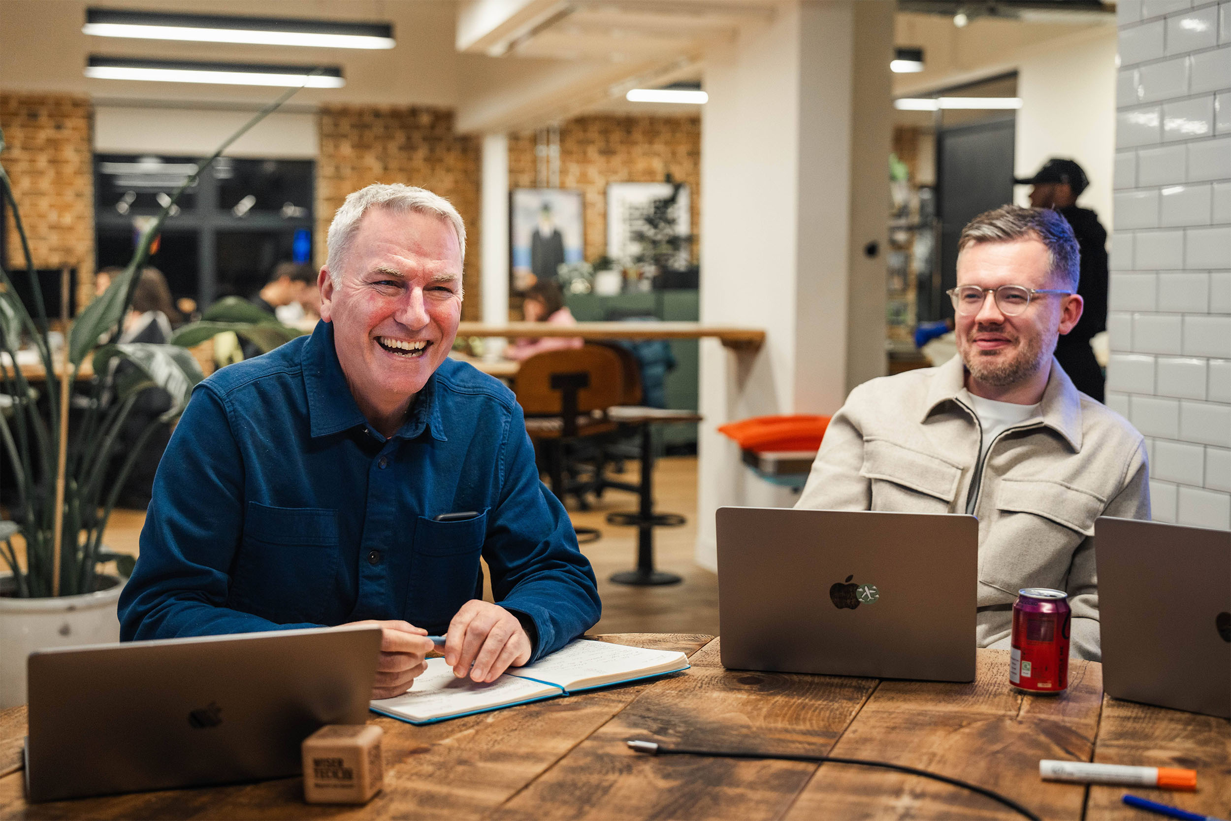 Two men, one in his thirties, one in his fifties, are having a meeting. Both smiling, one has a laptop open and the other is writing in a notebook