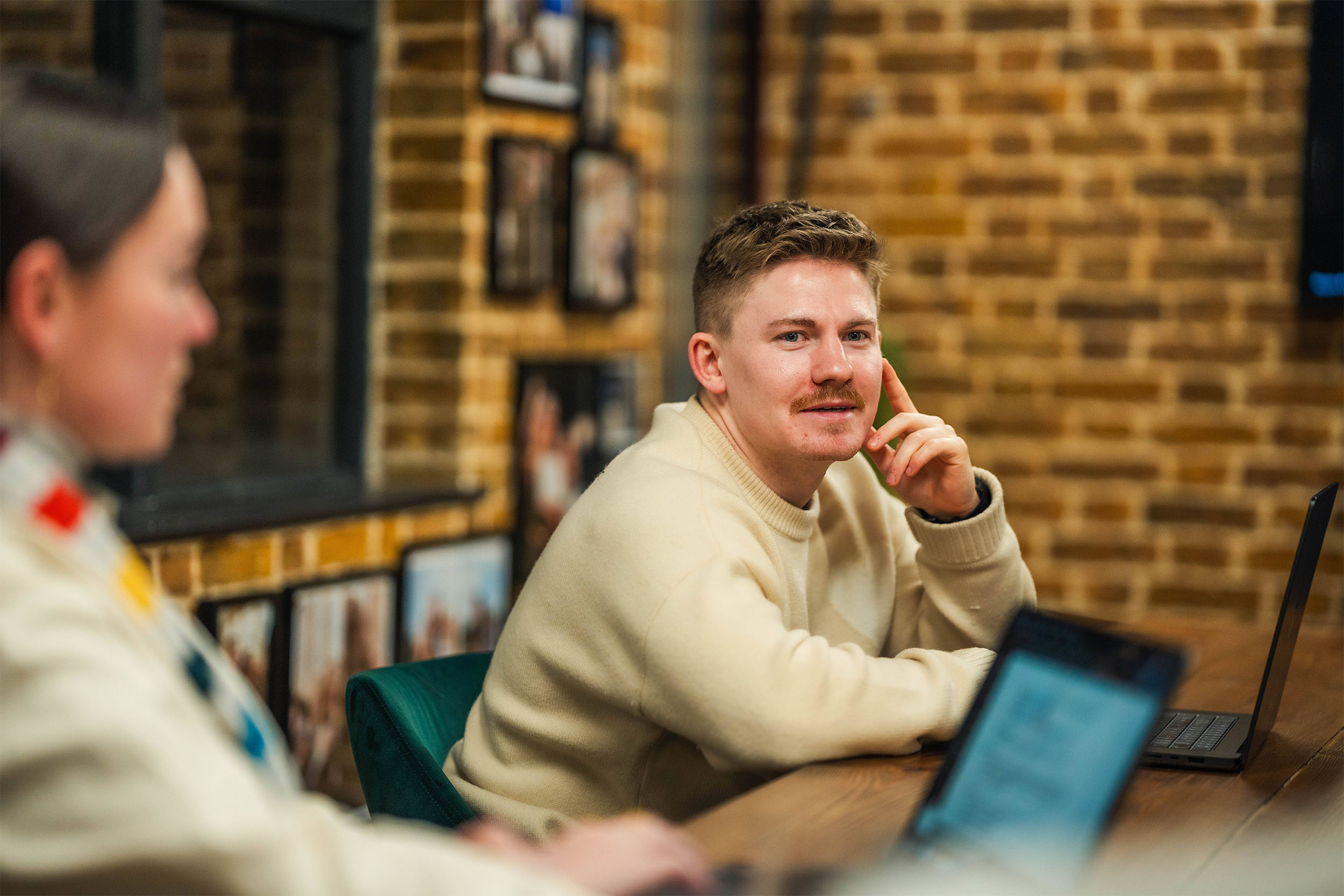 White blonde man with a moustache, wearing a cream knitted jumper, is speaking to somebody across the room