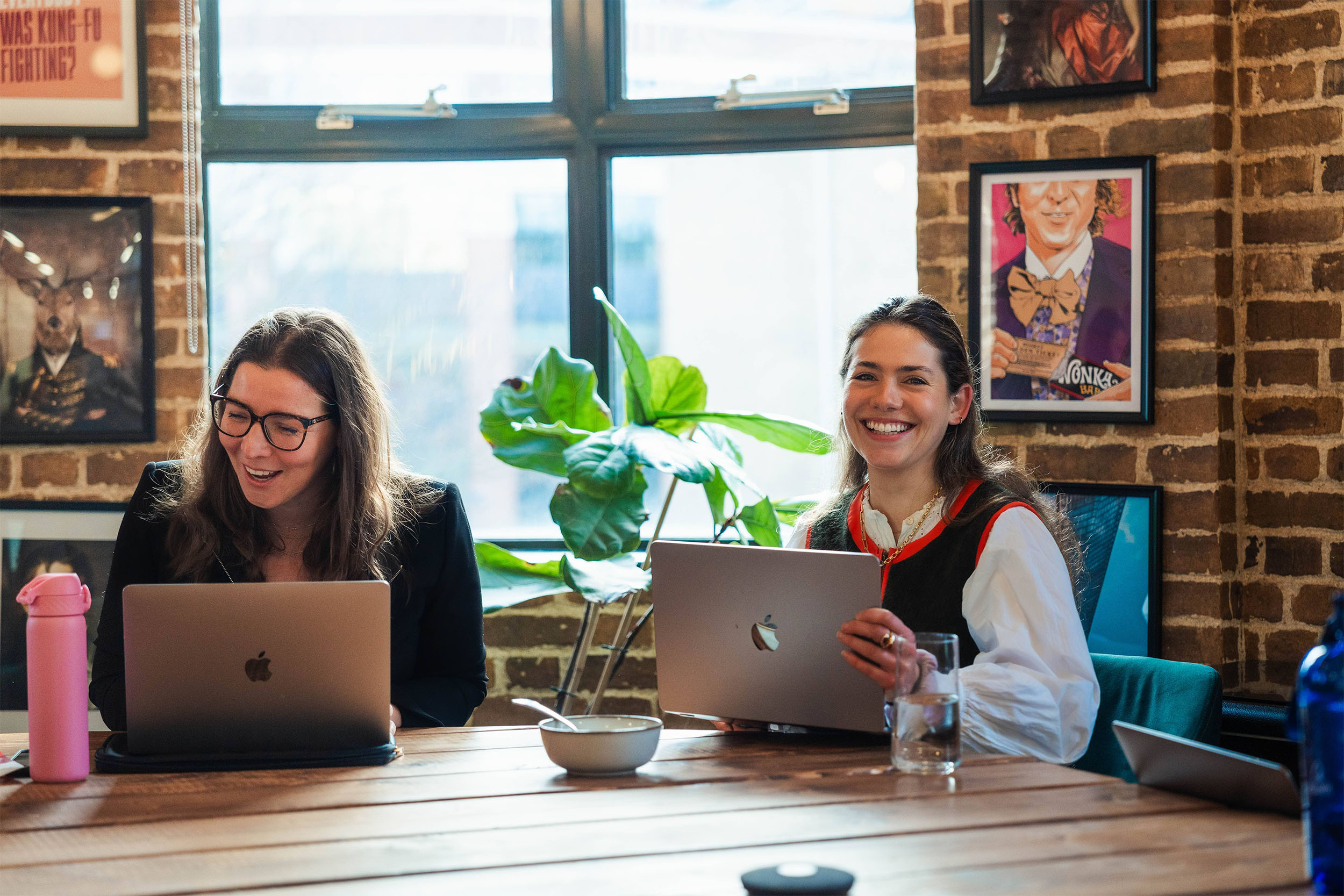Two women during a meeting with their laptops, both white with brown hair, one is looking at the camera and smiling, the other is talking to somebody out of frame about something on her laptop
