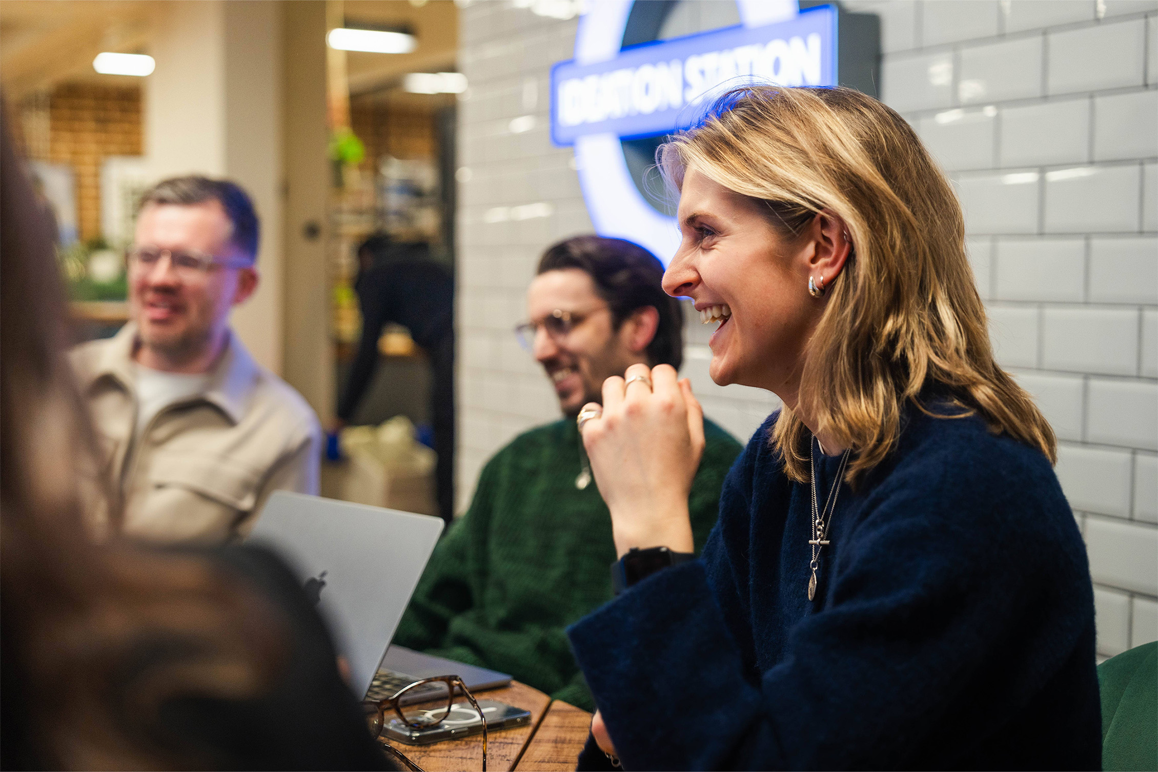 Close up of one woman, with 2 people out of focus behind her. She's smiling during a meeting, it's a side profile photo. She's blonde, wearing lots of necklaces and earrings