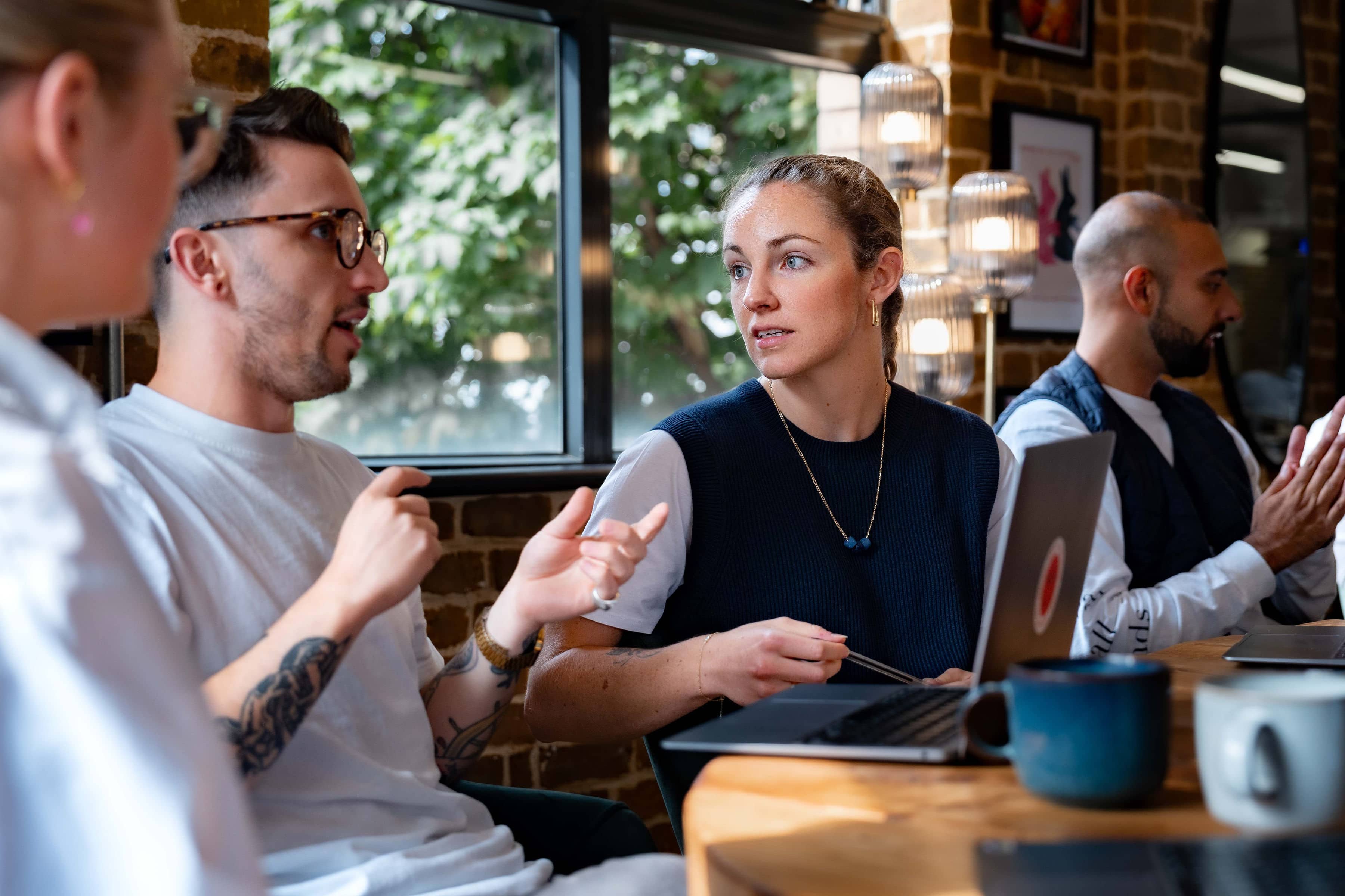 A busy meeting room, focusing in on two people. James is a white man with brown hair, glasses and arm tattoos, having a conversation with Emma, a white woman with her hair in one braid, wearing a white t-shirt with a navy vest on top