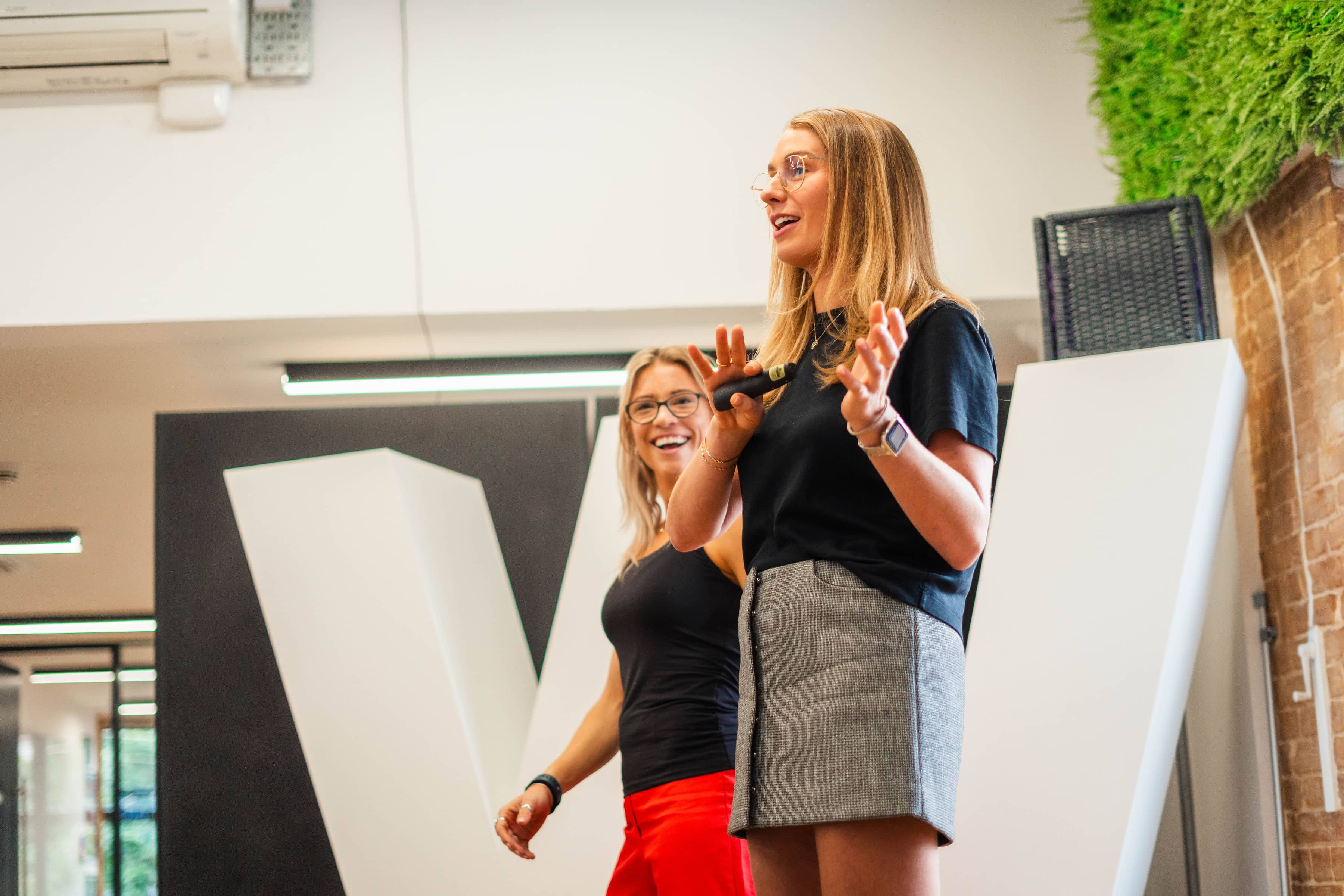 Head of People and COO delivering a talk. Both blonde white women. Both wearing black tops, one wears a grey skirt and the other red trousers