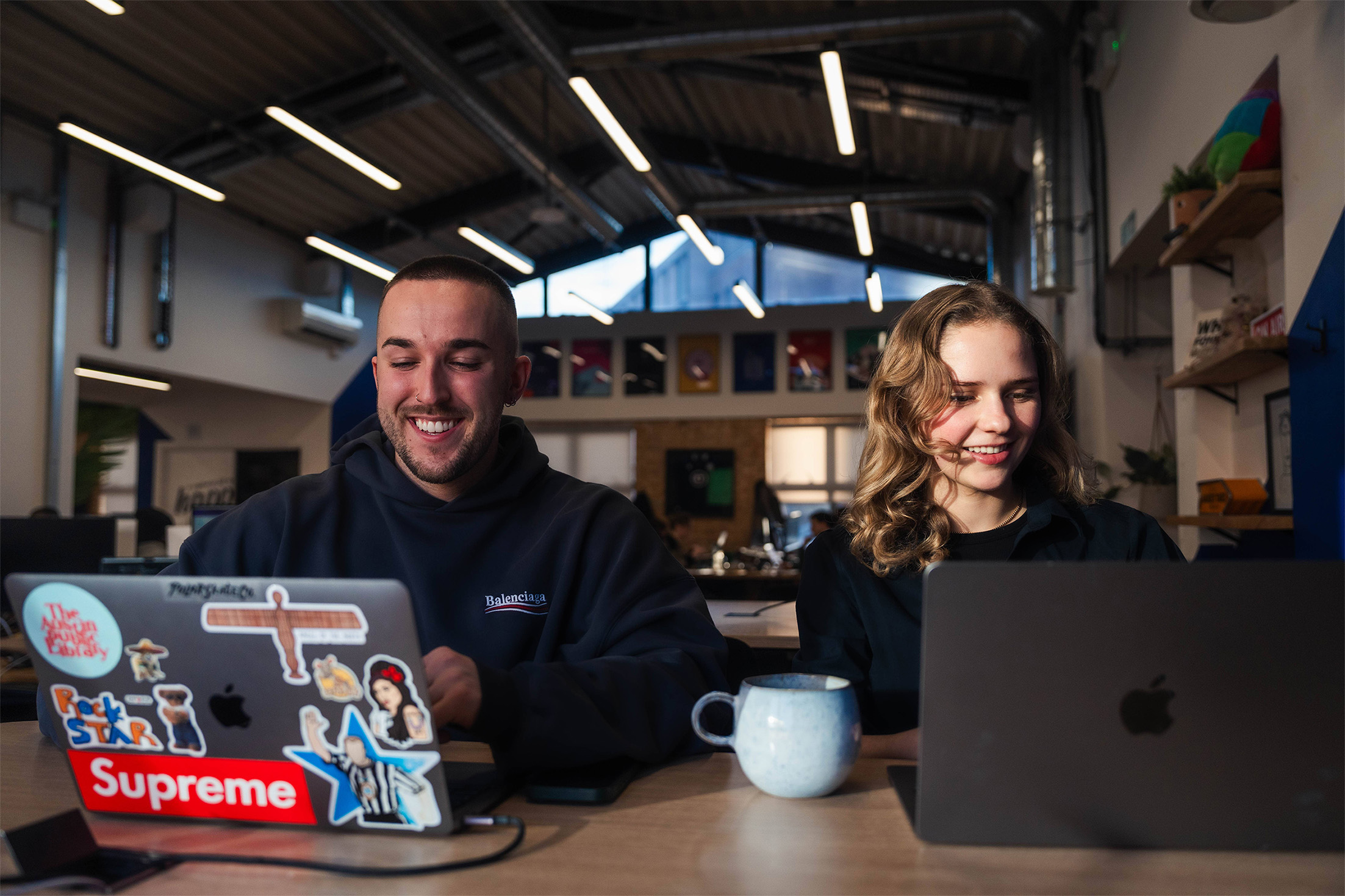 Two young adult collegues working together on laptops. Craig has short cropped hair and is wearing a dark hoodie, Eva has long dark blonde hair and is wearing a long sleeved dark top and is drinking a coffee