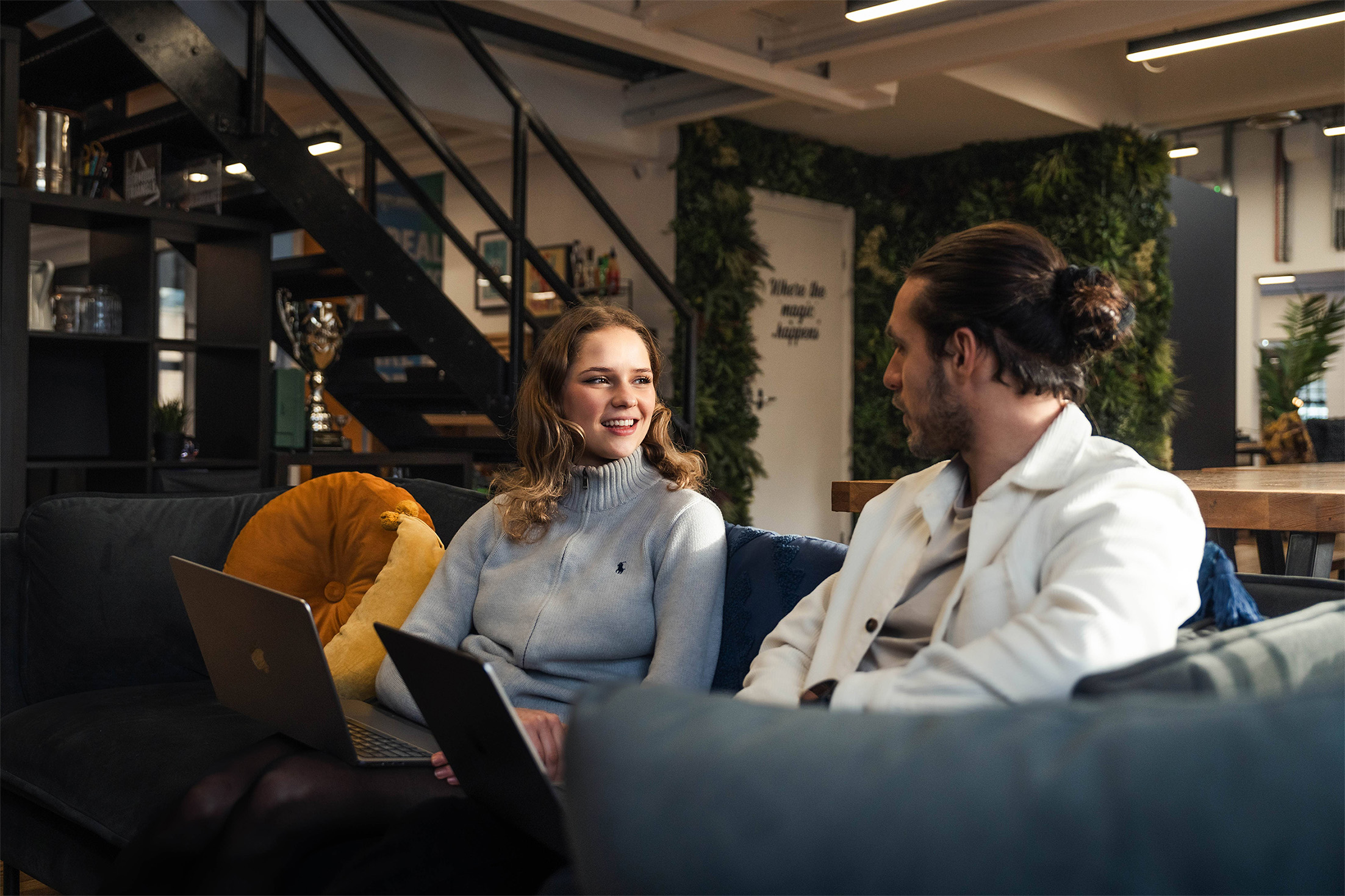 Our Development team working on a sofa together, they both have their laptops with them. Eva is wearing a grey zip up jumper, Charlie has his hair in a bun, and wears a white shirt